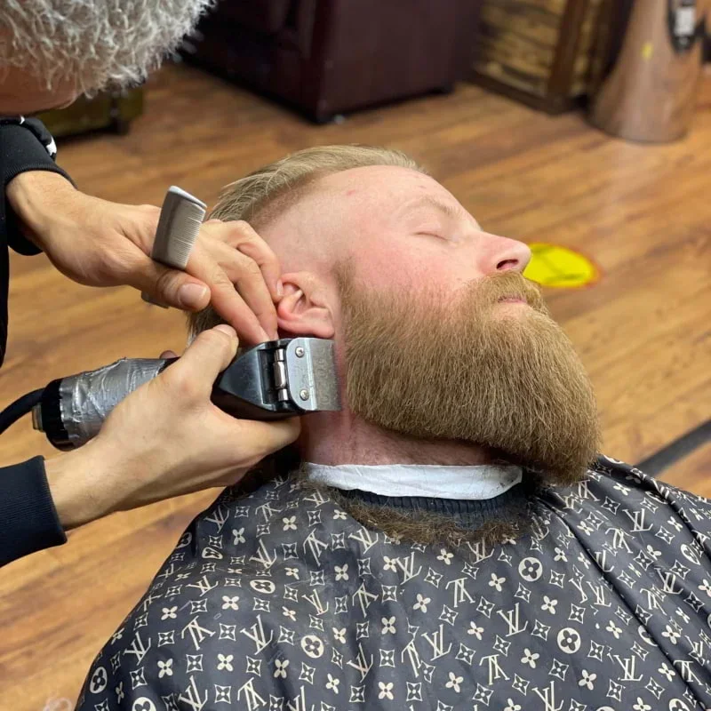 A man with a well-groomed beard is getting a haircut at a barbershop. The barber is using electric clippers on the side of his head while the man sits with his eyes closed, wearing a barbershop cape with a Louis Vuitton pattern.