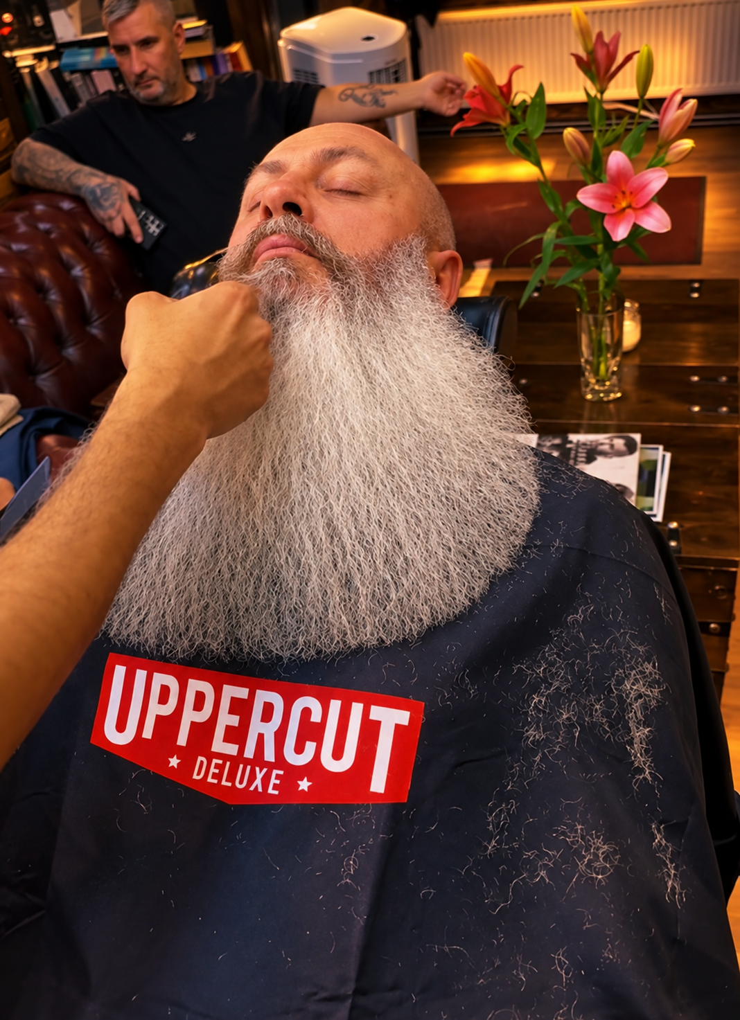 A man with a large white beard and closed eyes is getting a beard trim at a barbershop. The barber's hand is near his chin. Another man with tattoos on his arms is in the background, sitting on a leather chair, holding a remote. There is a vase with pink lilies and lily buds on the table, with some magazines and books around.