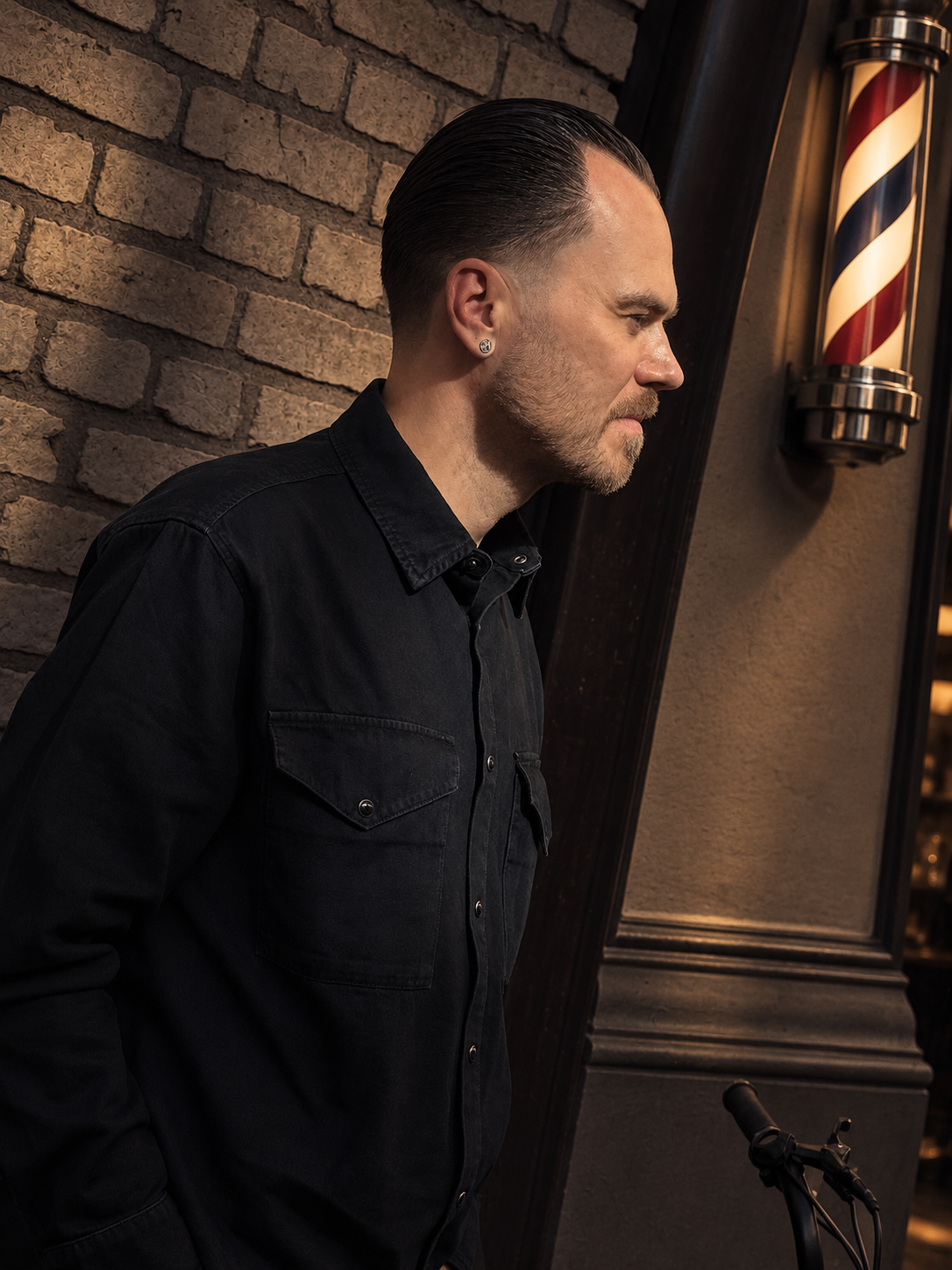 A man with slicked-back hair and a beard standing indoors against a brick wall, wearing a black shirt, near a barber's pole.