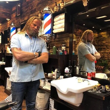 Man with long hair and beard standing in barber shop, looking into mirror with arms crossed, surrounded by barber tools and products.