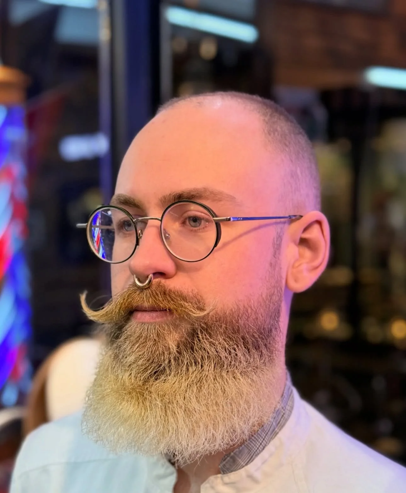 A man with a shaved head, round glasses, a septum piercing, and a thick, well-groomed beard. He is in a dimly lit indoor setting, possibly a bar or restaurant.