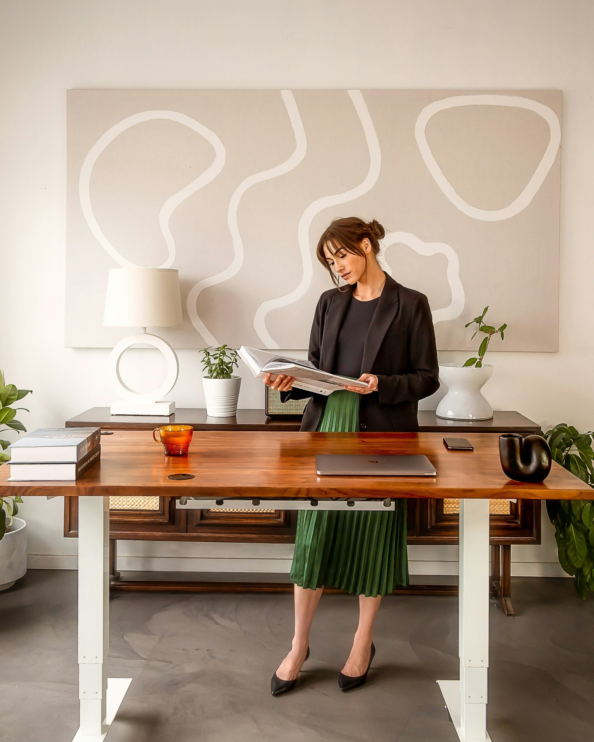 red haired woman in green dress, standing at desk, looking at a book