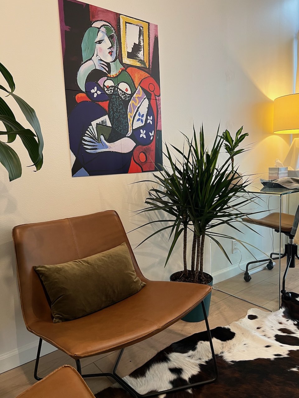 Living room corner with a brown leather chair, dark green cushion, a potted plant, colorful abstract artwork on the wall, a desk with a chair, a lamp, and a cowhide rug.
