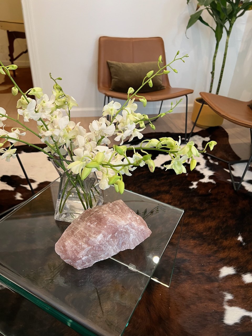 A floral arrangement of white orchids in a glass vase on a glass table, with a large pink salt lamp, in a living room with a cowhide rug, and chairs with leather cushions and a green potted plant in the background.