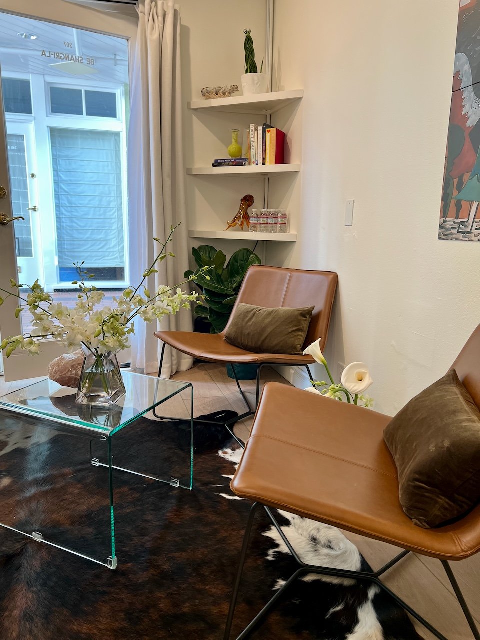 A cozy living room corner with tan leather chairs, a glass coffee table with a flower arrangement, a cowhide rug, and a potted plant on the floor. A built-in white wall shelf holds books, a yellow vase, and decorative items. A sliding glass door with white curtains leads outside.