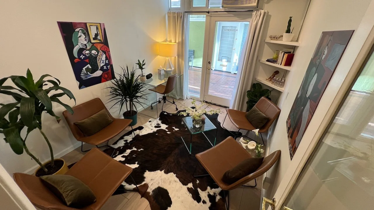 Living room with four brown leather chairs around a cowhide rug, glass coffee table with white flowers, potted plants, artwork on the walls, a glass desk with a brown chair, and double glass doors leading outside.