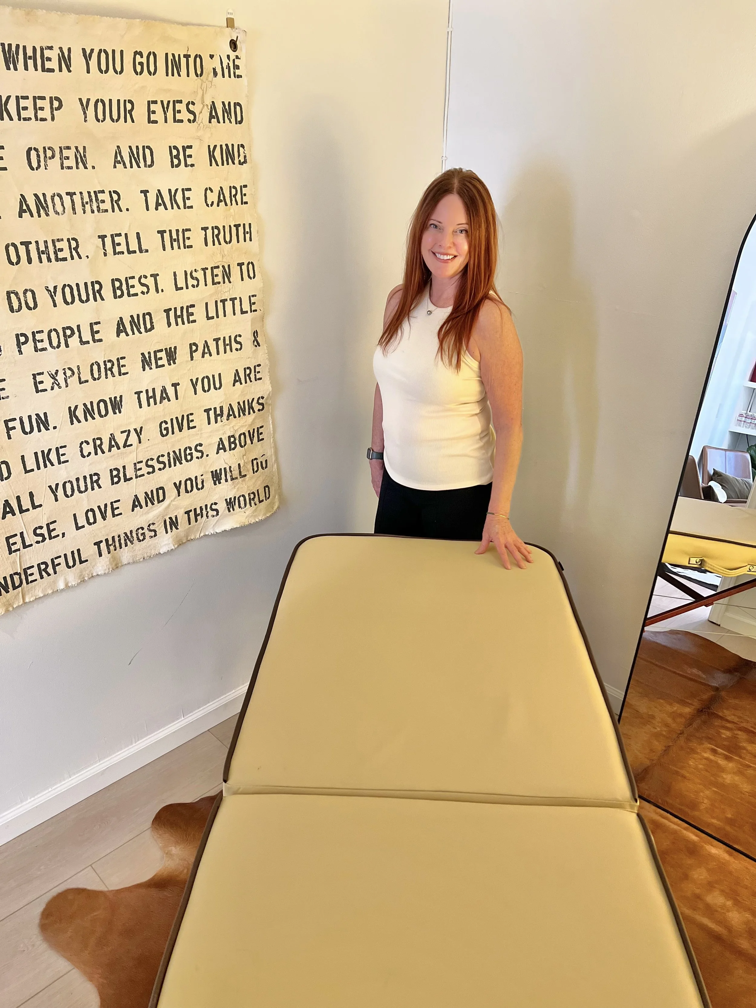 A smiling woman with long red hair standing behind a yellow table, in a room with a cream-colored wall, a large mirror, and a cream-colored and brown animal-patterned floor rug. A framed quote on fabric hangs on the wall.