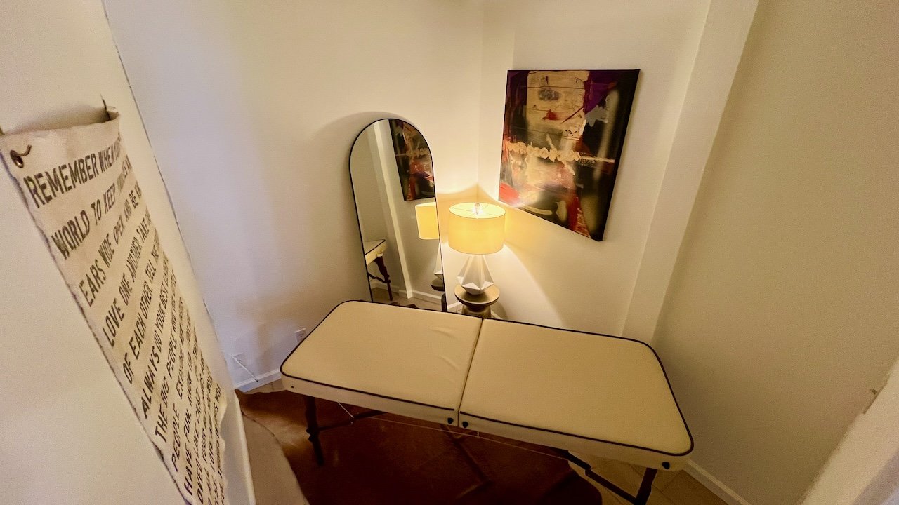 A small treatment room with a white adjustable massage bed, a tall mirror, a table lamp on a small side table, and a colorful abstract painting on the wall.