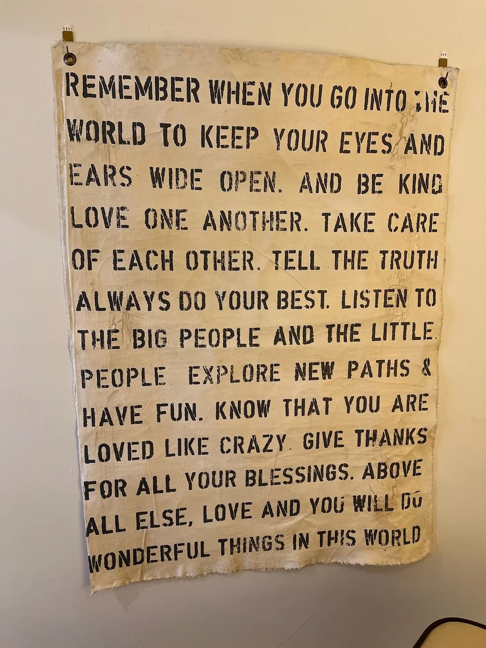 A beige wall hanging with a positive message printed in bold black letters. The message encourages kindness, truth, listening, exploring new paths, loving, giving thanks, and appreciating blessings.