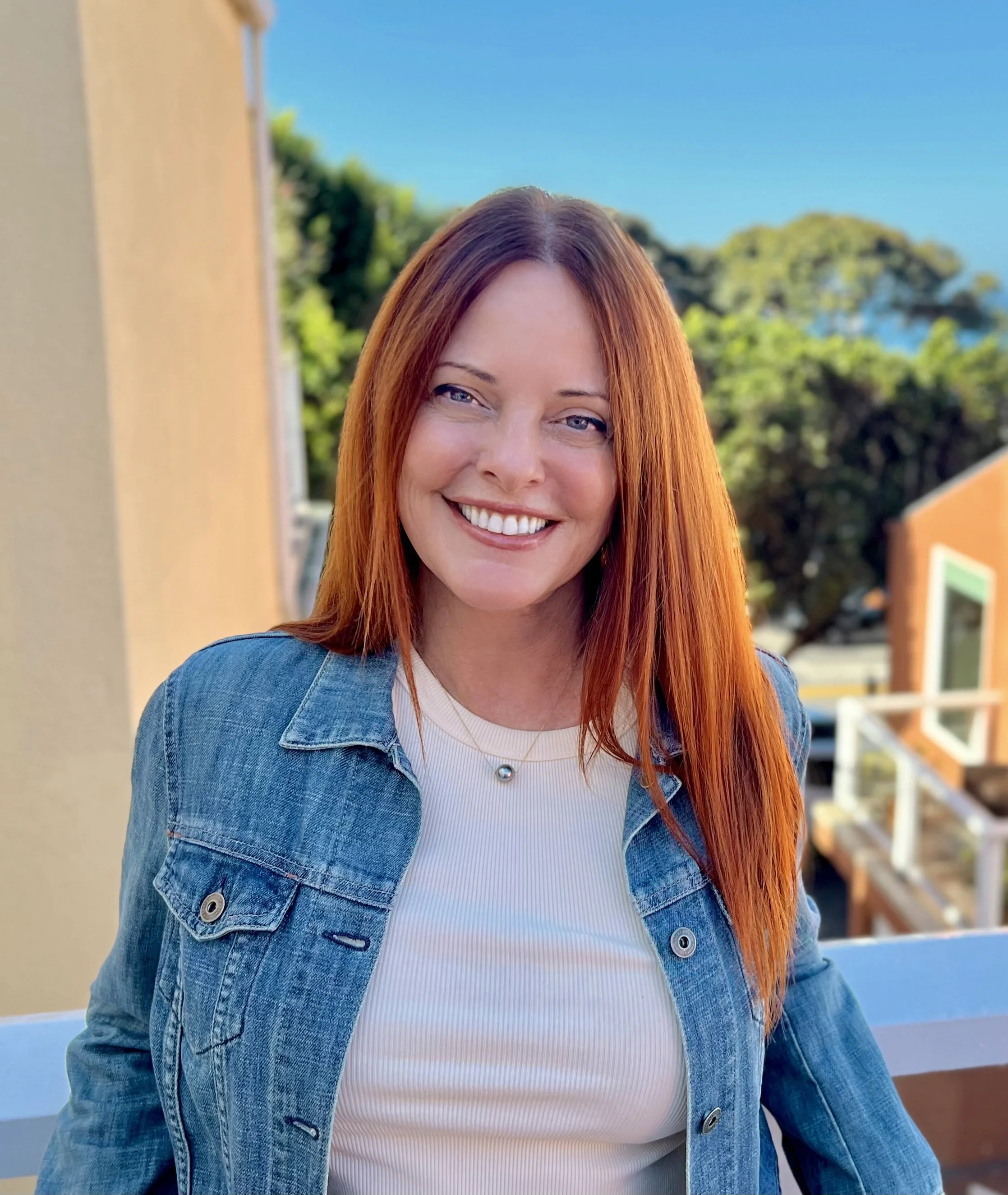 A woman with long red hair smiling outdoors, wearing a denim jacket and a white shirt, with trees and a blue sky in the background.