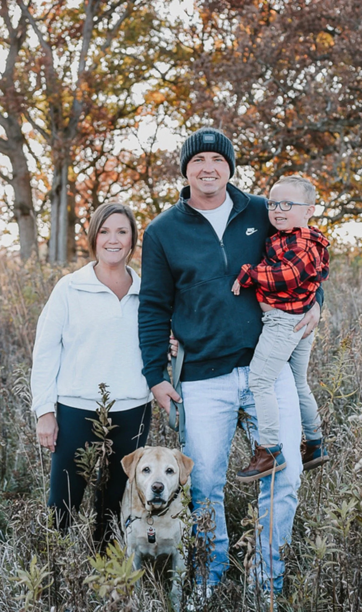 A family of three with a dog standing outdoors in a field during autumn, with colorful fall leaves on trees in the background. The woman is smiling and standing to the left, the man is holding a young boy in a red plaid jacket on the right, and the dog is sitting on the ground in front of them.