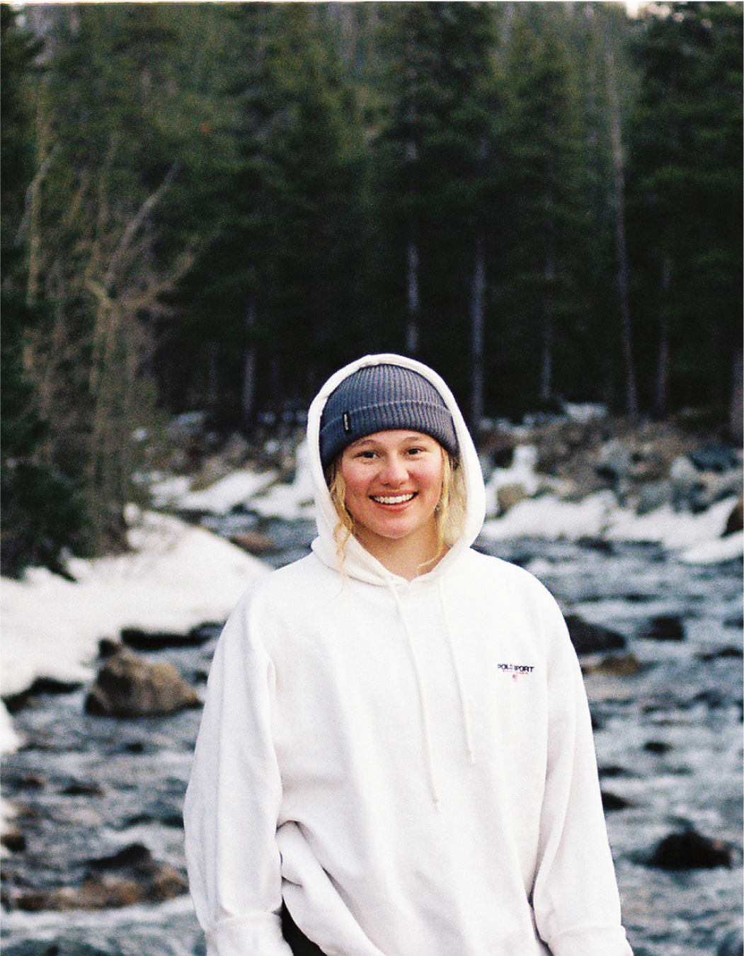 A woman in a white hoodie and a gray beanie standing near a river with rocks and snow in a forested area.