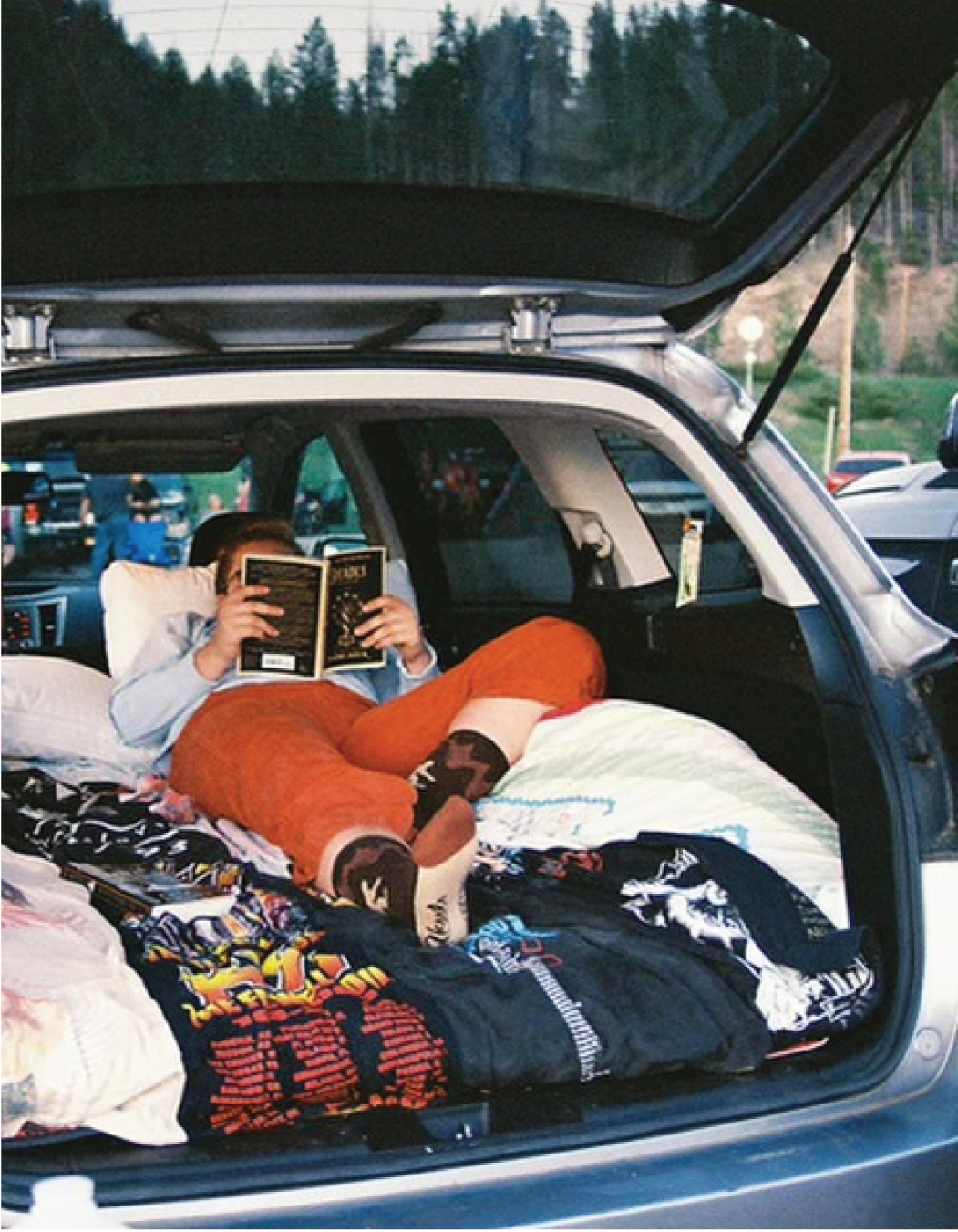 A person relaxing in the back of a car with the trunk open, lying on a bed surrounded by blankets and pillows, reading a book.