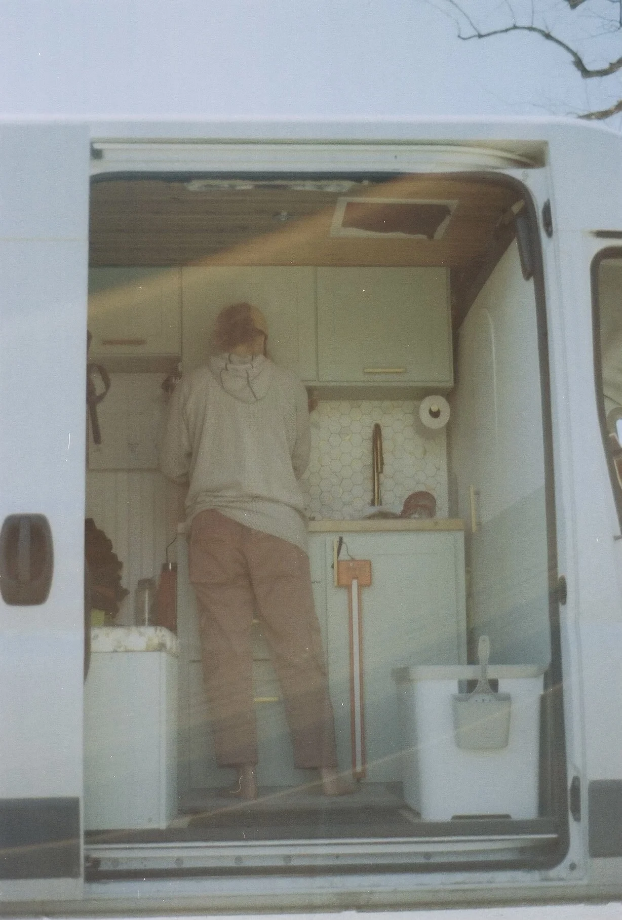 Person standing inside a tiny camper kitchen, viewed through the open door, with cabinets, a sink, paper towel roll, and various items visible.
