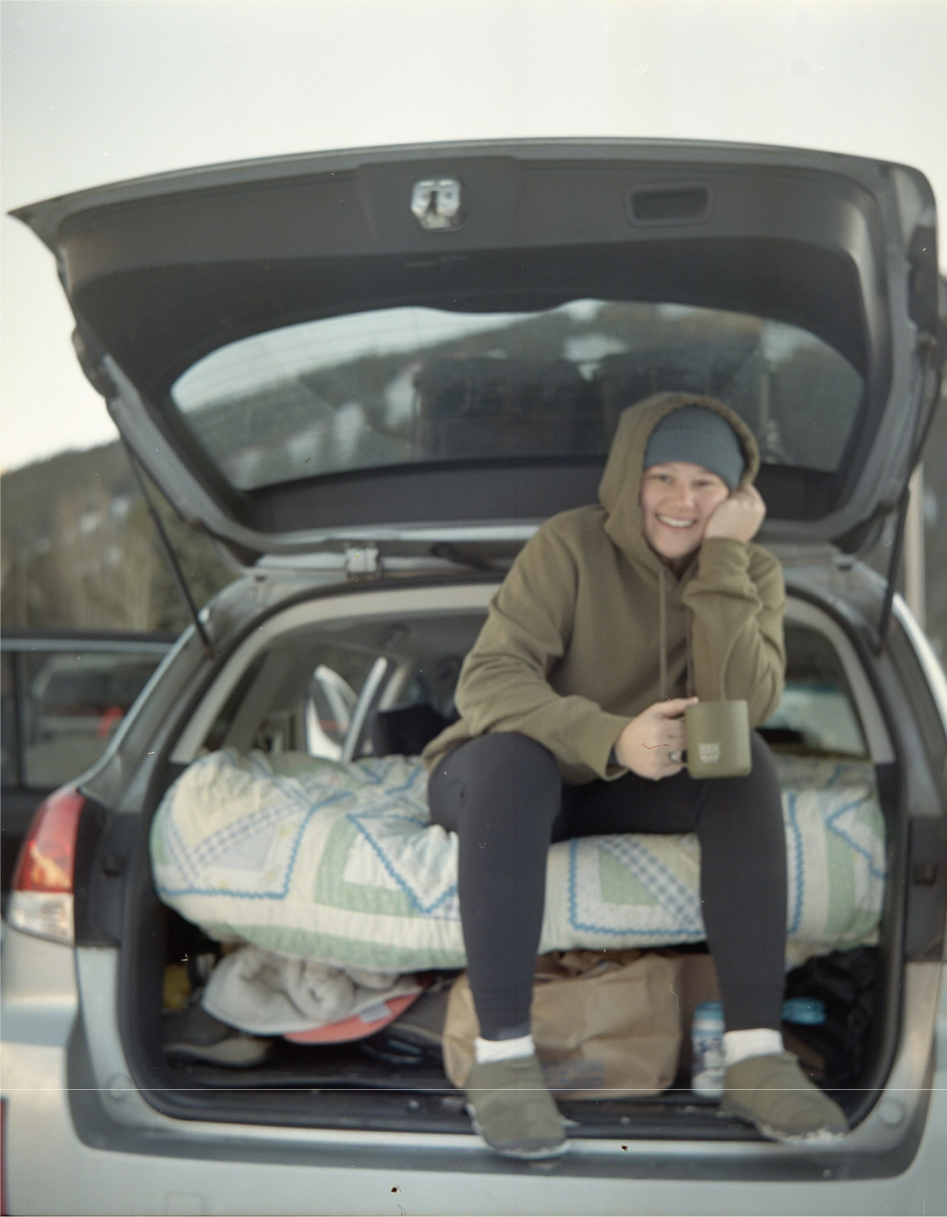 A person sitting in the open trunk of a car, smiling, wearing a gray beanie and a brown hoodie, holding a mug, with camping gear and blankets inside the trunk, outdoors in a natural setting.