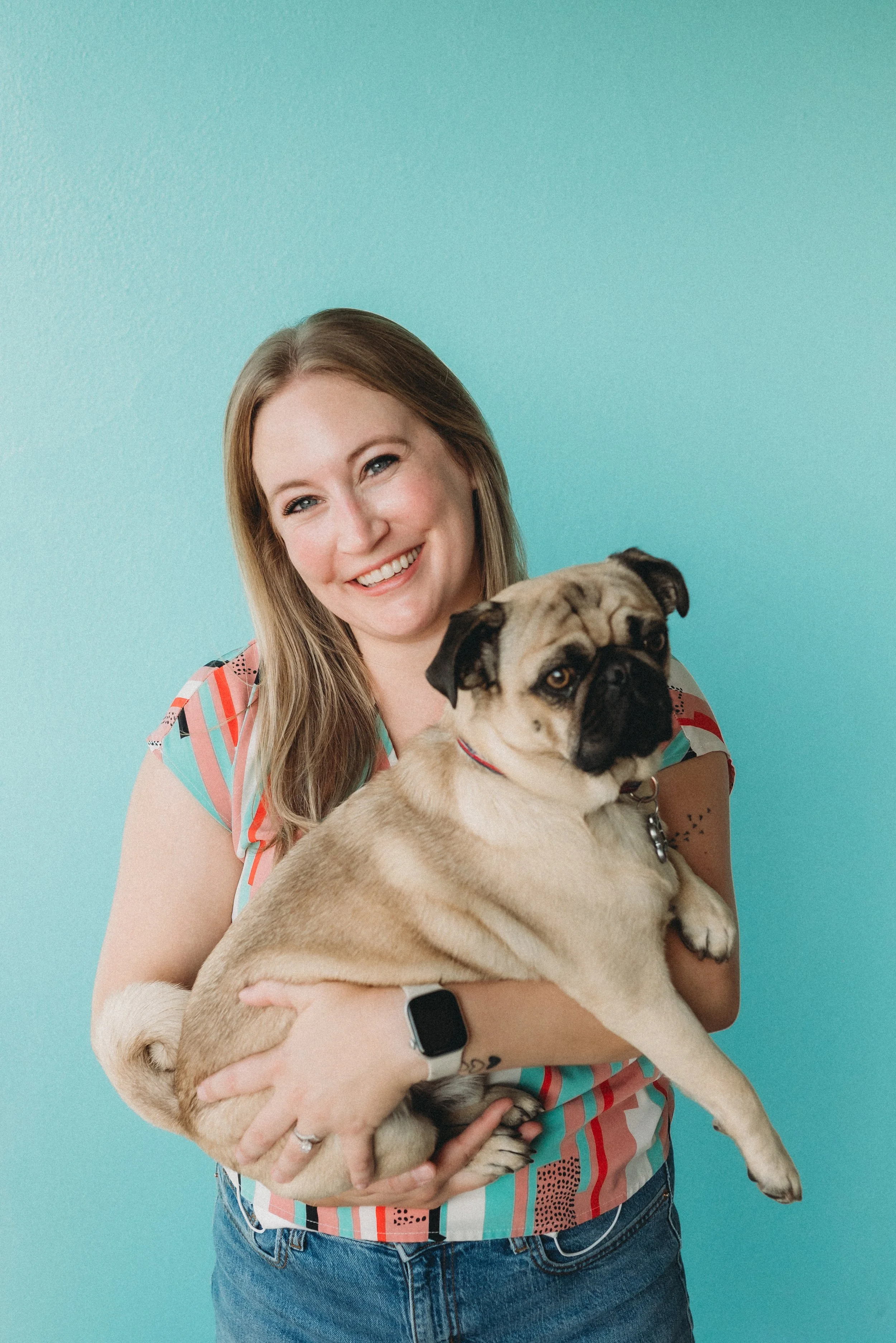 A woman with long, light brown hair smiling and holding a large, light-colored pug dog against a turquoise background. The woman is wearing a colorful short-sleeve shirt and a smartwatch.