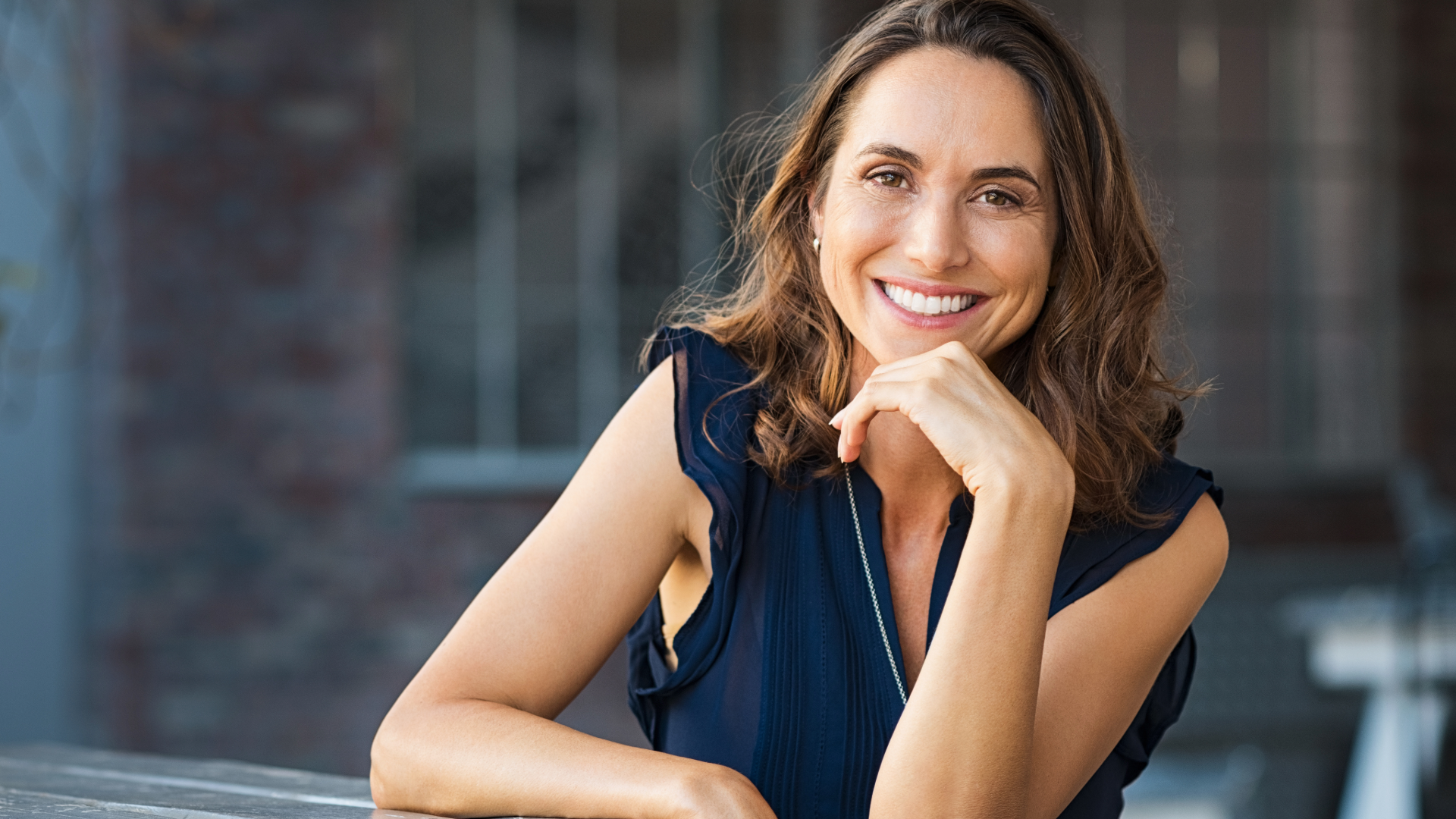 A smiling woman with wavy brown hair, wearing a navy blue sleeveless top, resting her chin on her hand and looking at the camera outdoors.