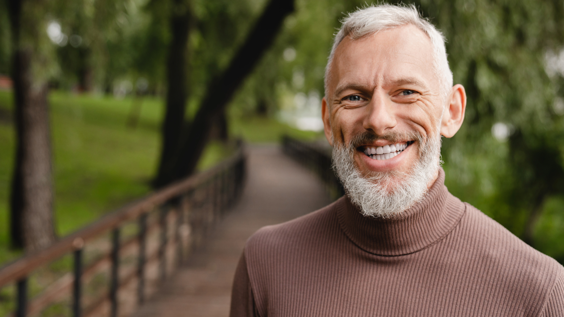 A smiling elderly man with a white beard and short gray hair standing on a park pathway with green trees and fencing in the background.