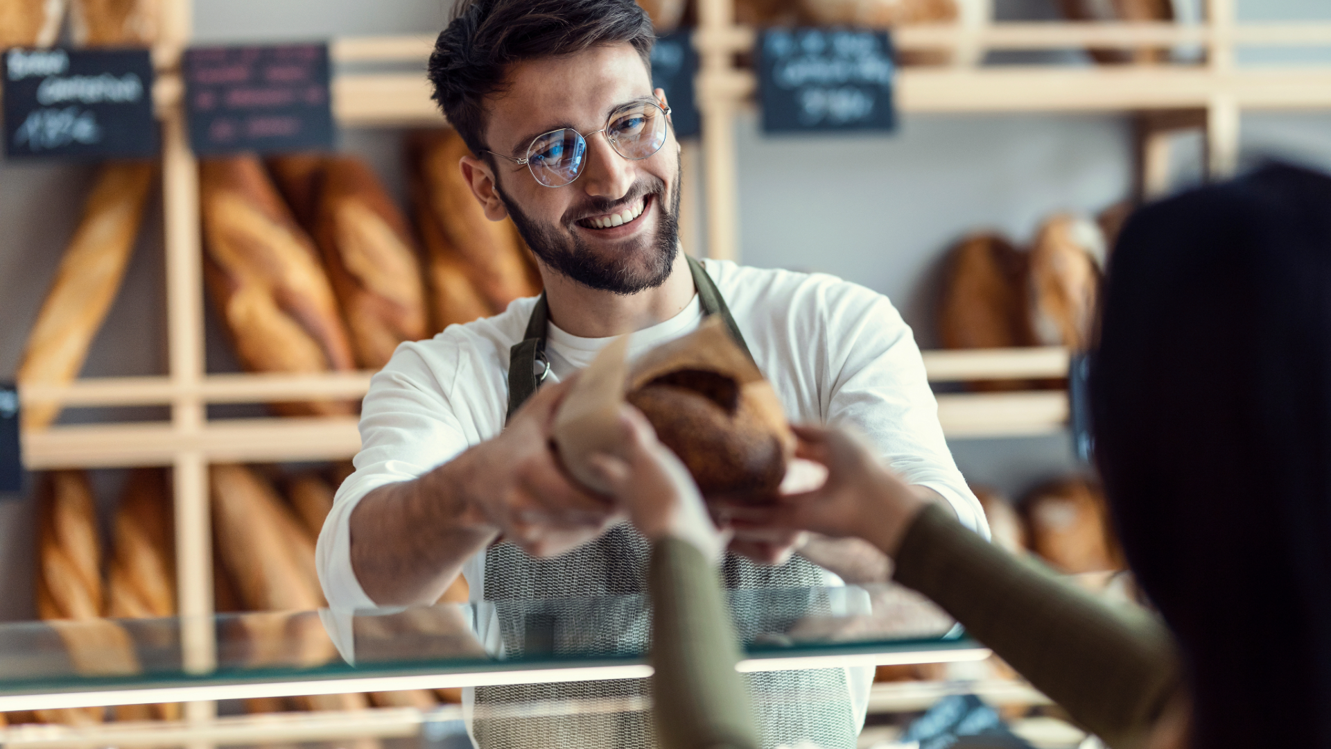 A smiling man in glasses and an apron hands a loaf of bread to a customer in a bakery. Shelves with baguettes are in the background.
