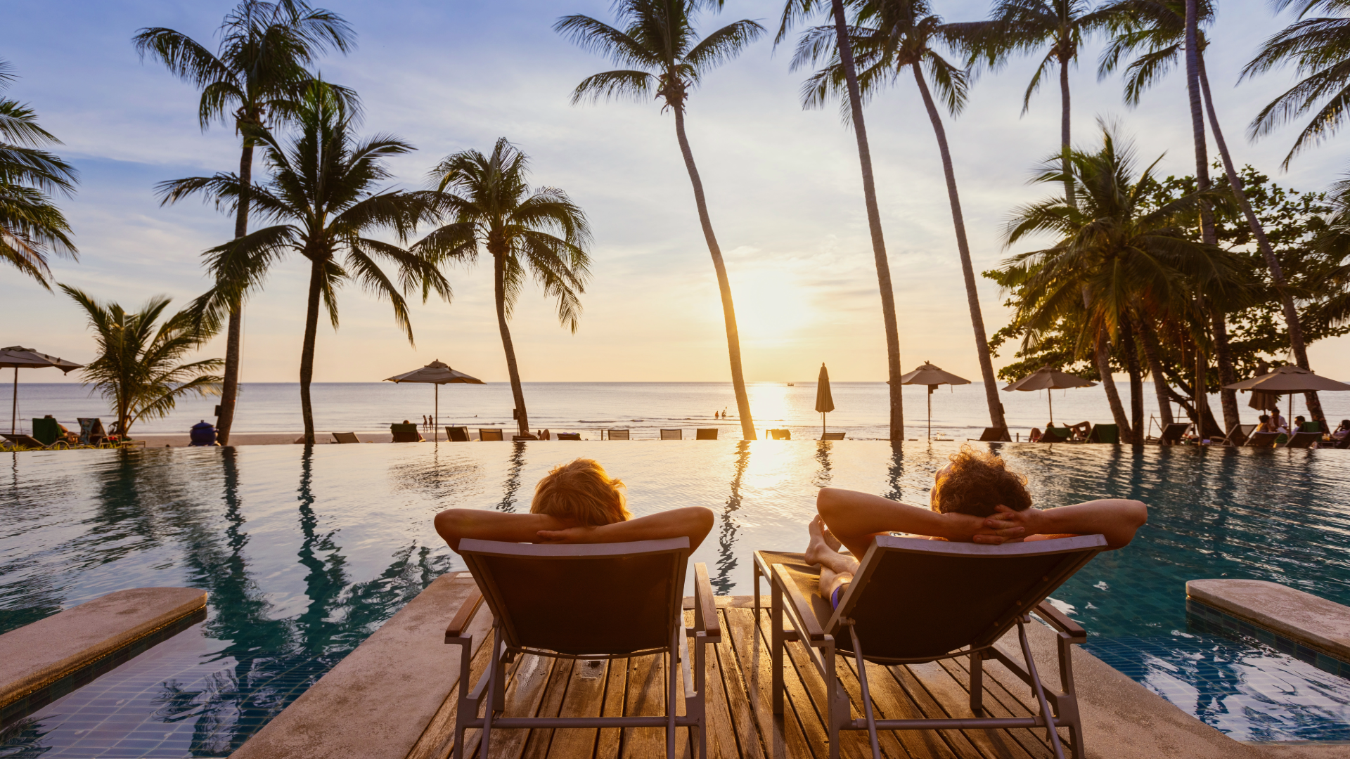 Two people relaxing on lounge chairs by a pool, overlooking a beach with palm trees and umbrellas at sunset.