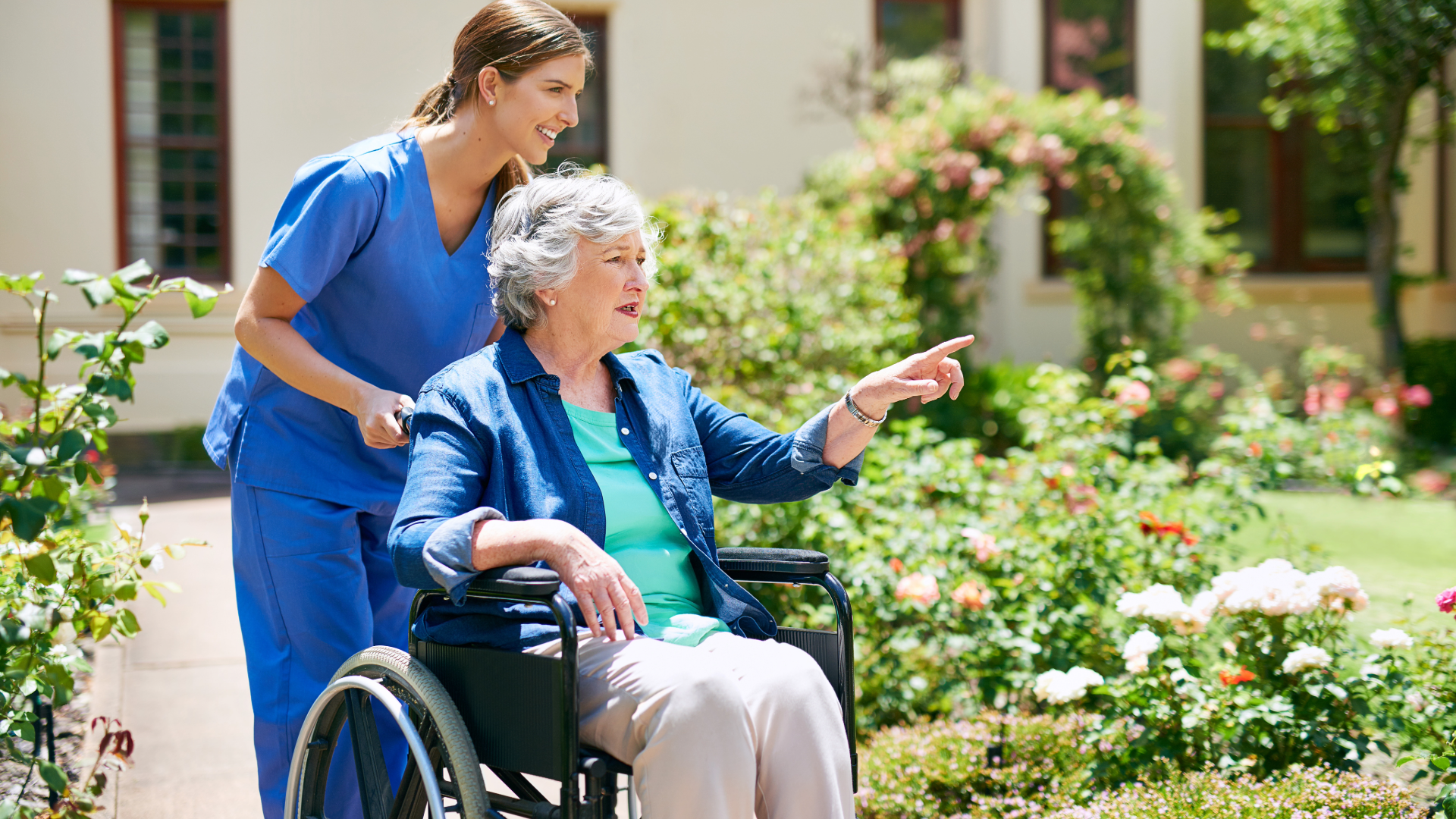 A healthcare worker in blue scrubs pushing an elderly woman in a wheelchair through a garden filled with blooming flowers and lush greenery.