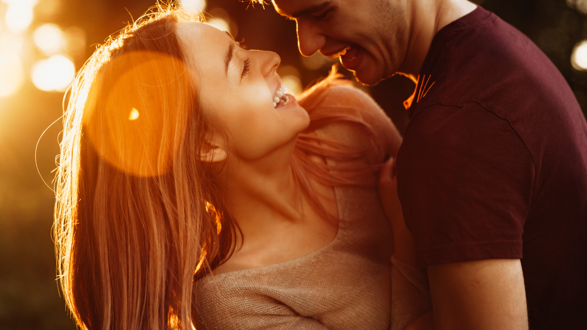 A young woman and a young man are close together, smiling and laughing as they look at each other. The woman has long red hair and the man is wearing a burgundy shirt. They are outdoors at sunset, with warm, golden light illuminating their faces and hair.