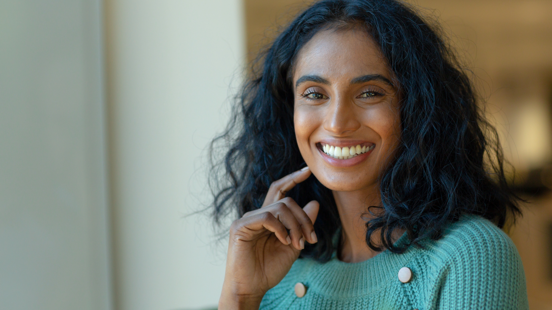 Close-up of a smiling woman with curly black hair wearing a green sweater, indoors with neutral background.