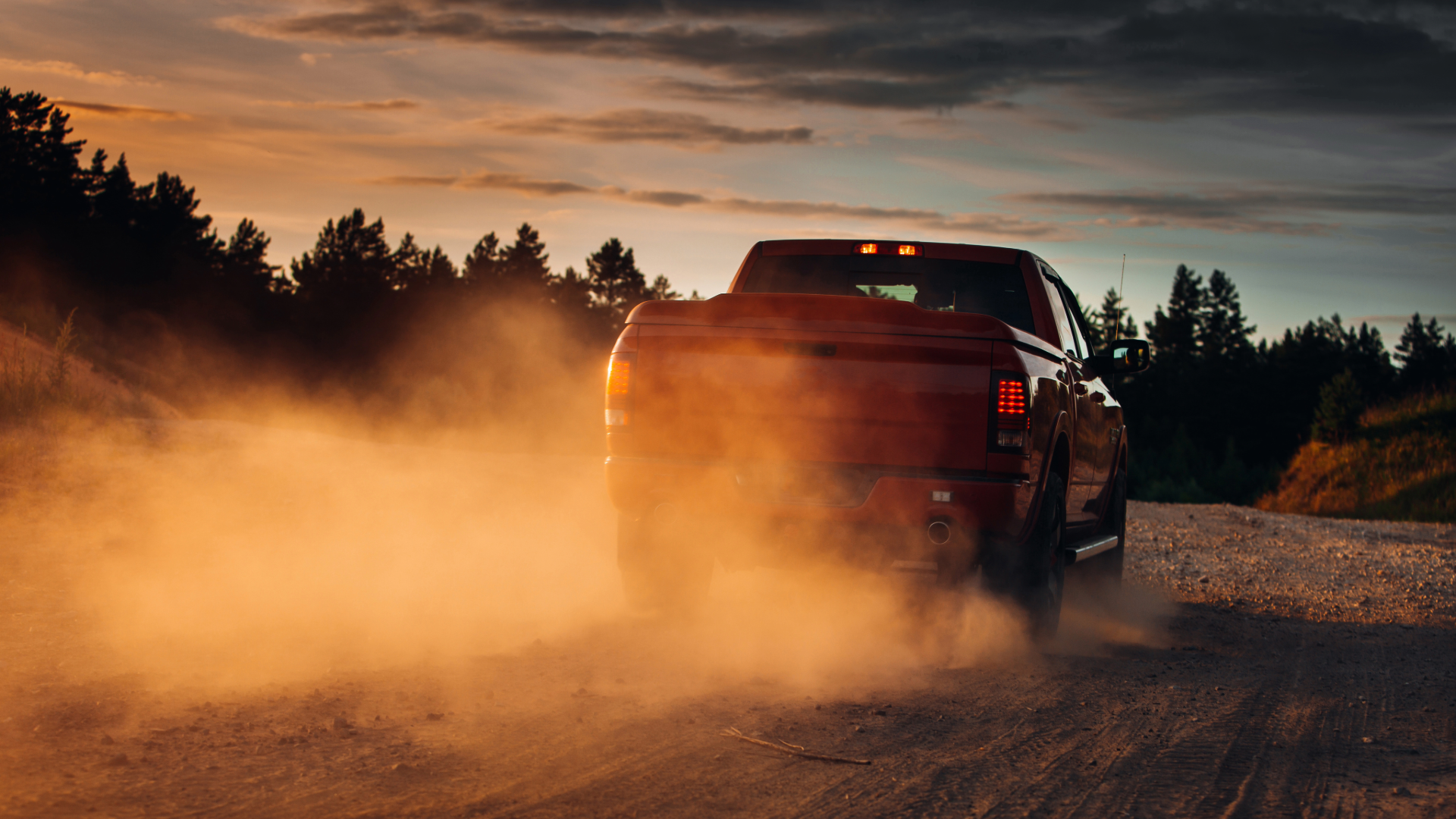 A red pickup truck driving on a dirt road at sunset, kicking up dust behind it with a forested landscape in the background.