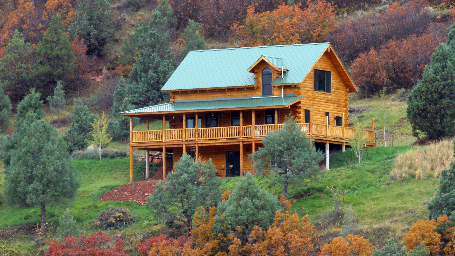 A wooden house with a metal roof on a hillside, surrounded by trees with autumn foliage.