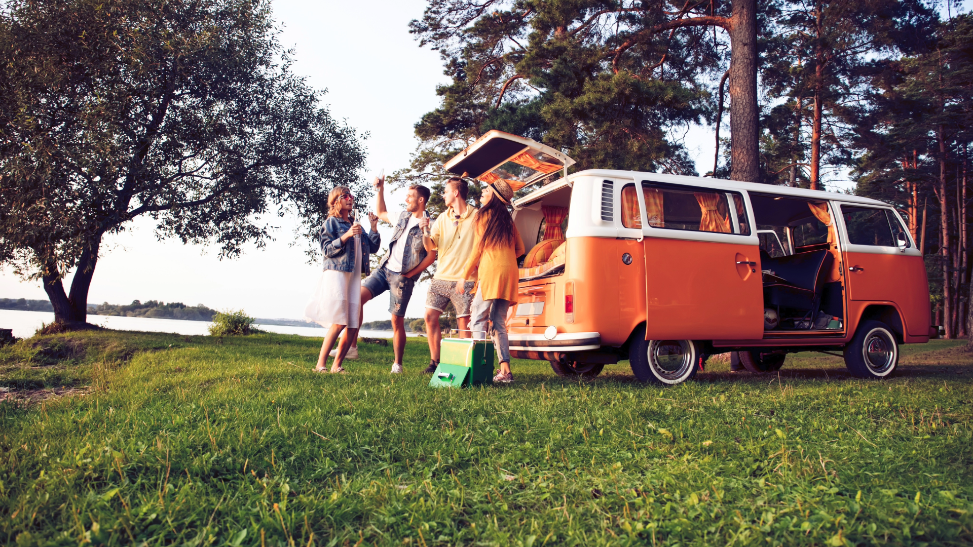Group of four friends enjoying a picnic outdoors near a lake, with a vintage orange and white van parked on the grass and tall trees in the background.