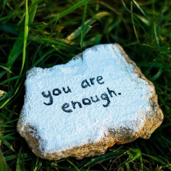 A painted rock with the words "you are enough" written on it in black ink, resting on green grass.