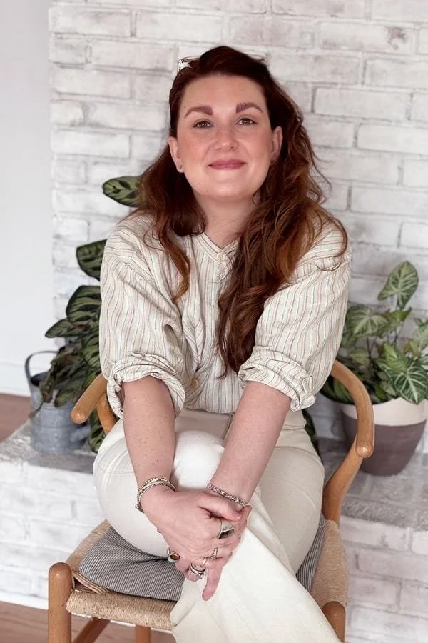 Dani-Rae Strohminger sitting indoors against a white brick wall with potted plants behind her, wearing a beige striped shirt and white pants.