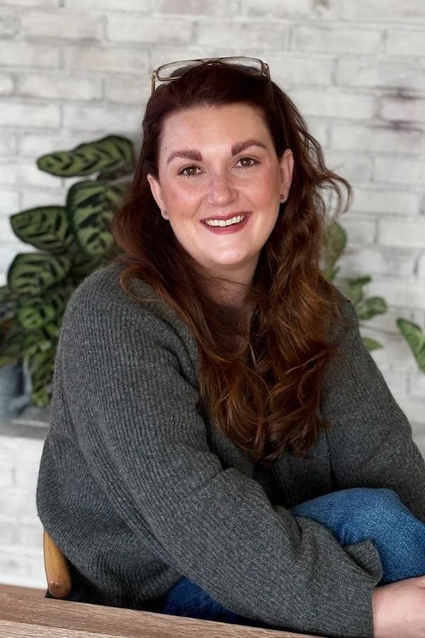 Dani-Rae Strohminger wearing glasses on her head and a gray sweater, smiling at the camera, with green plants and a white brick wall in the background.