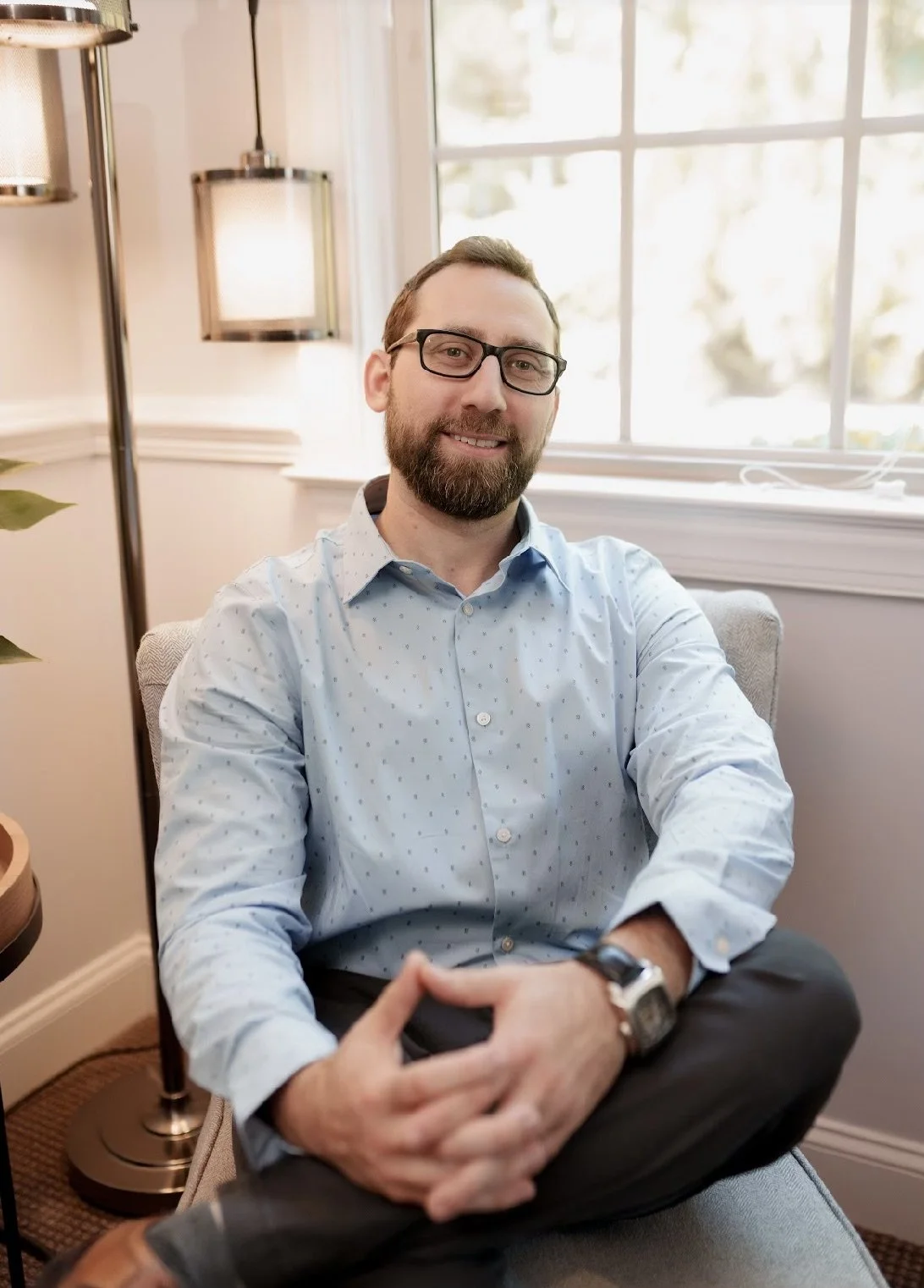 Man sitting in an armchair with hands clasped, wearing glasses and a light blue shirt, indoors by a window and lamp.