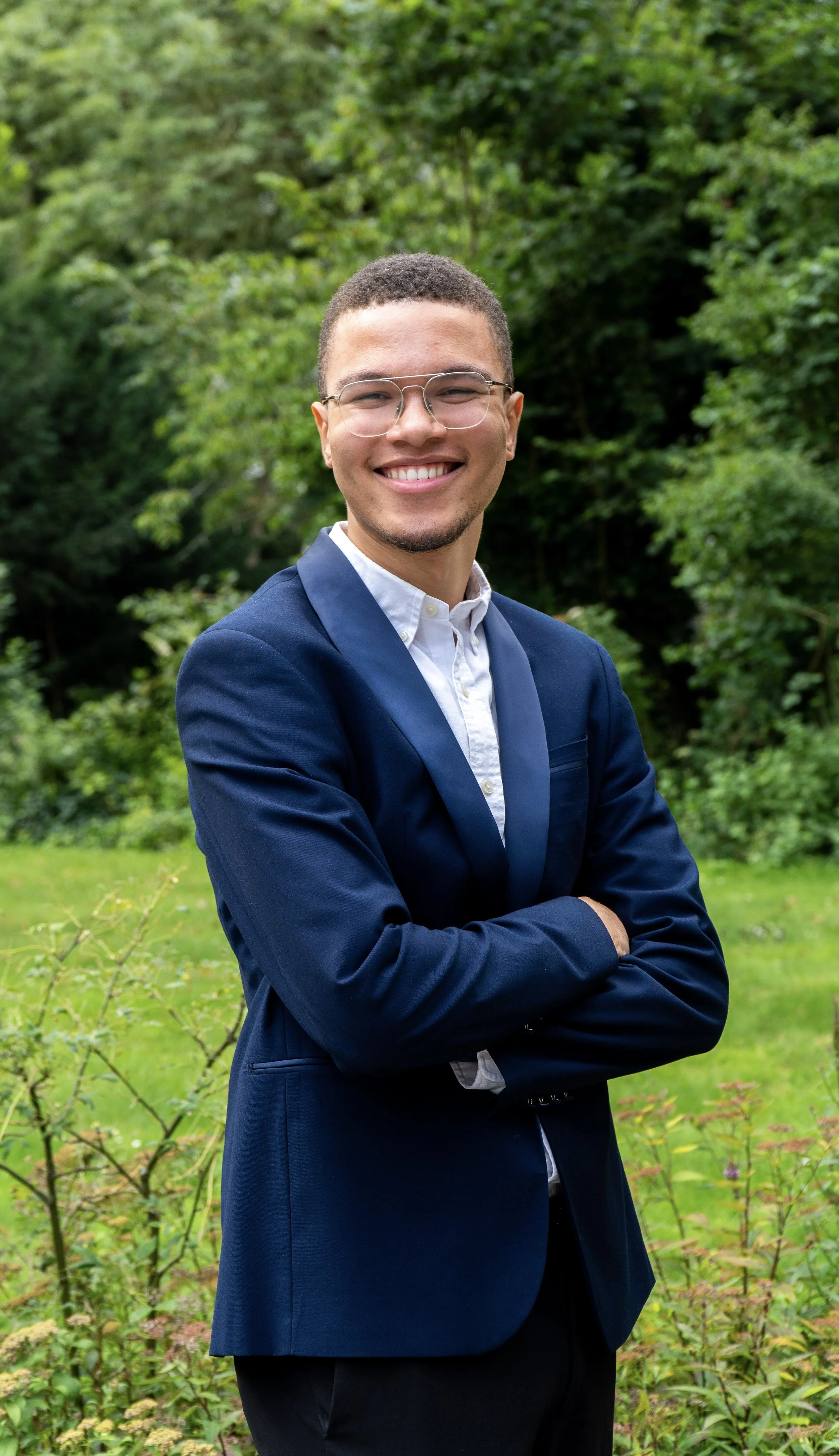 Lachende jonge man in formele kleding met bril, achtergrond van groen bos of park.