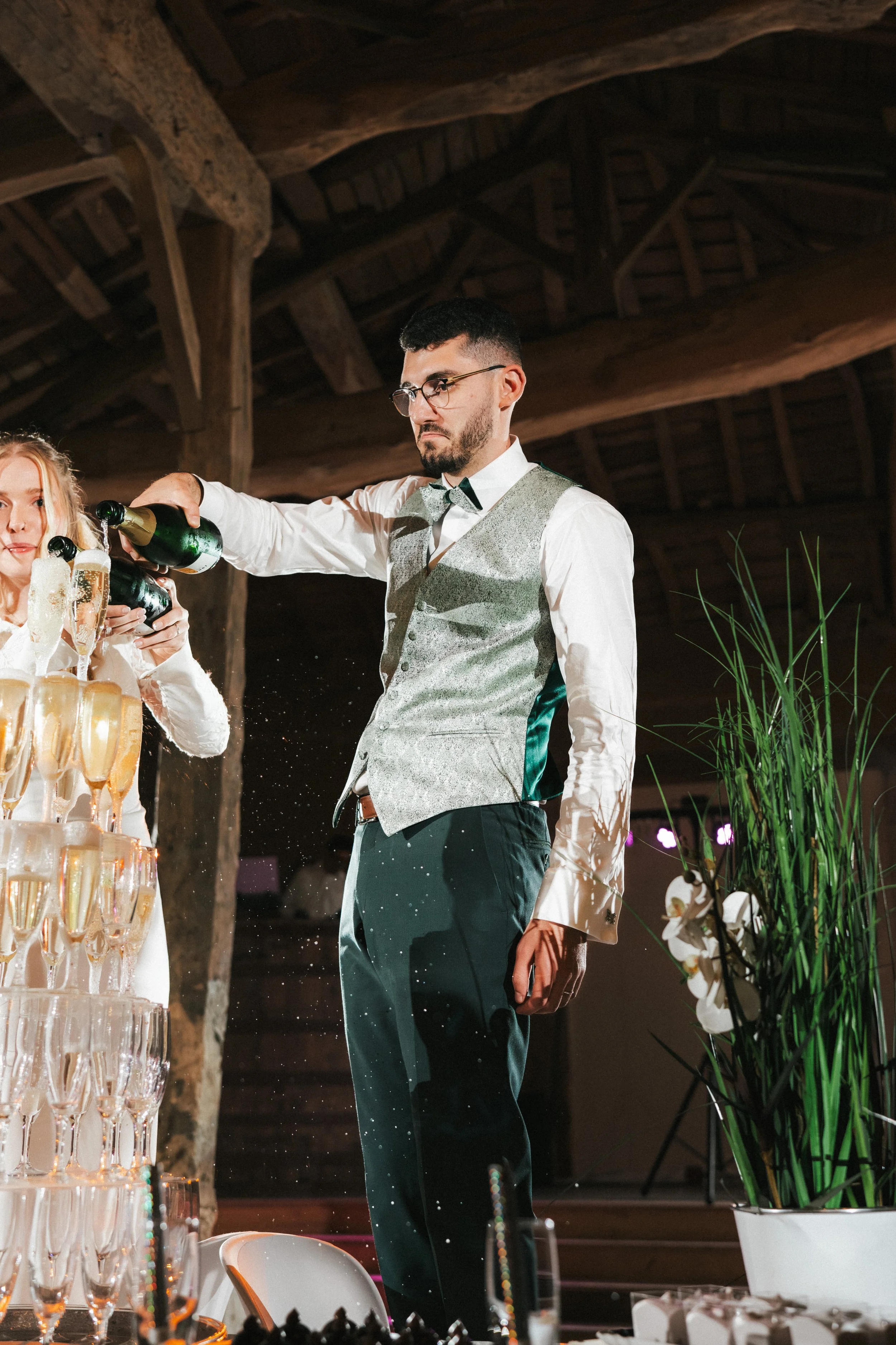 Un homme habillé en veston et chemise blanche, portant des lunettes, verse du champagne dans des flûtes lors d'une célébration dans une salle en bois. À côté, une femme tient plusieurs bouteilles de champagne.