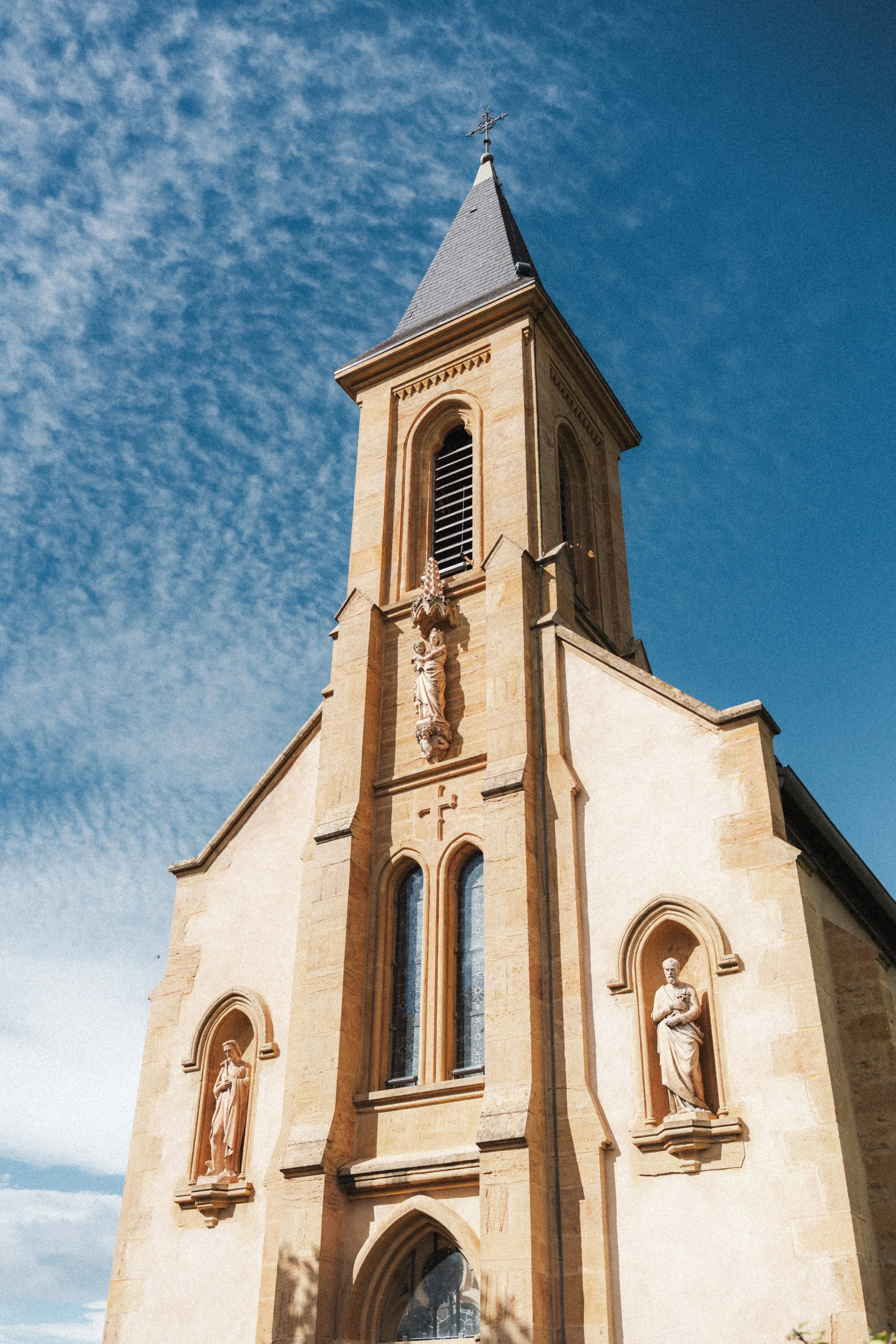 Une église en pierre avec des statues religieuses et un clocher pointu sous un ciel bleu avec des nuages dispersés.