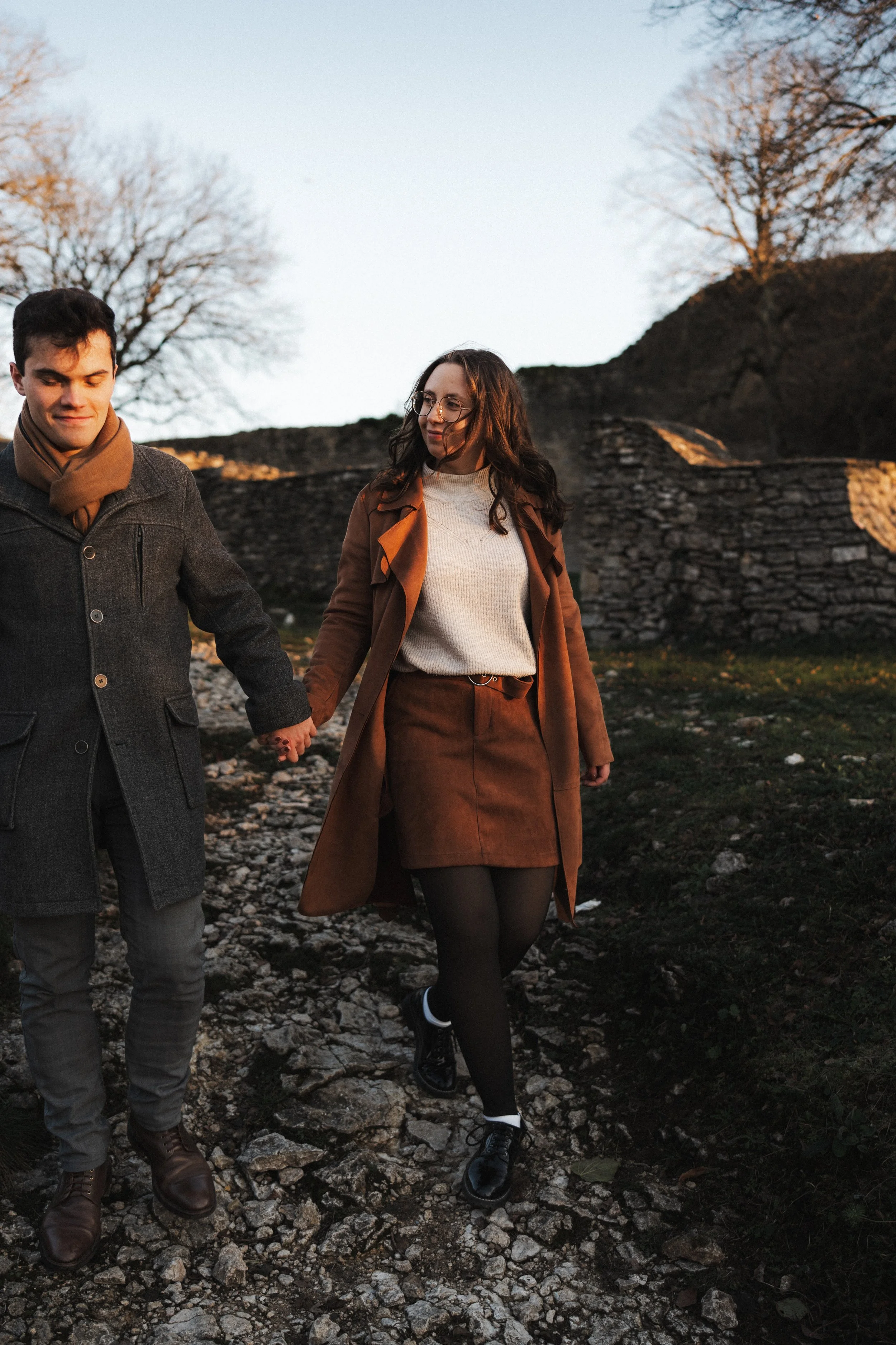 Un couple marchant main dans la main sur un chemin rocheux en plein air, avec des arbres dénudés et un mur en pierre en arrière-plan, au coucher du soleil.