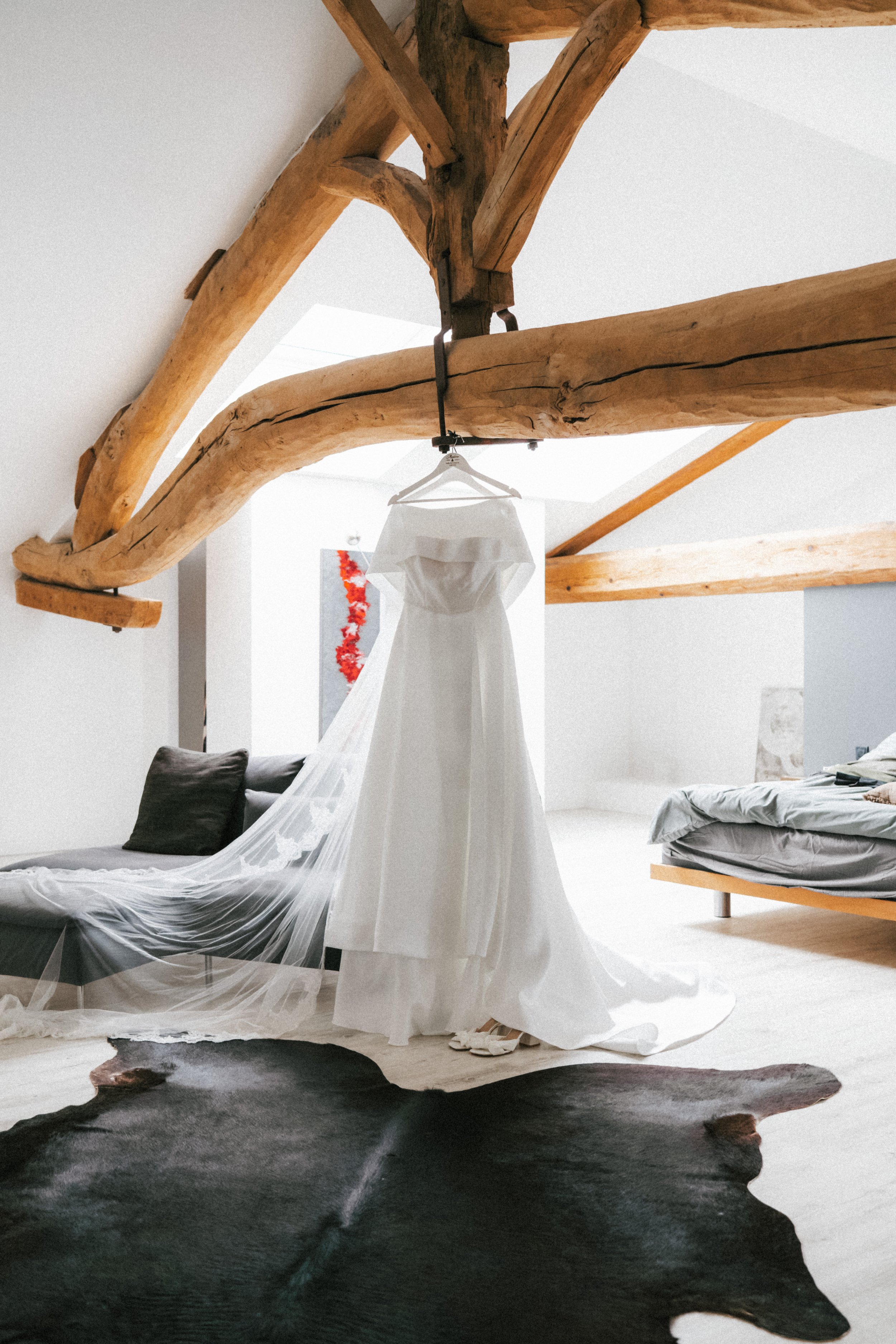 Une robe de mariage blanche suspendue, dans une pièce avec poutres en bois apparentes, un canapé gris avec coussins et un lit avec une couverture grise.