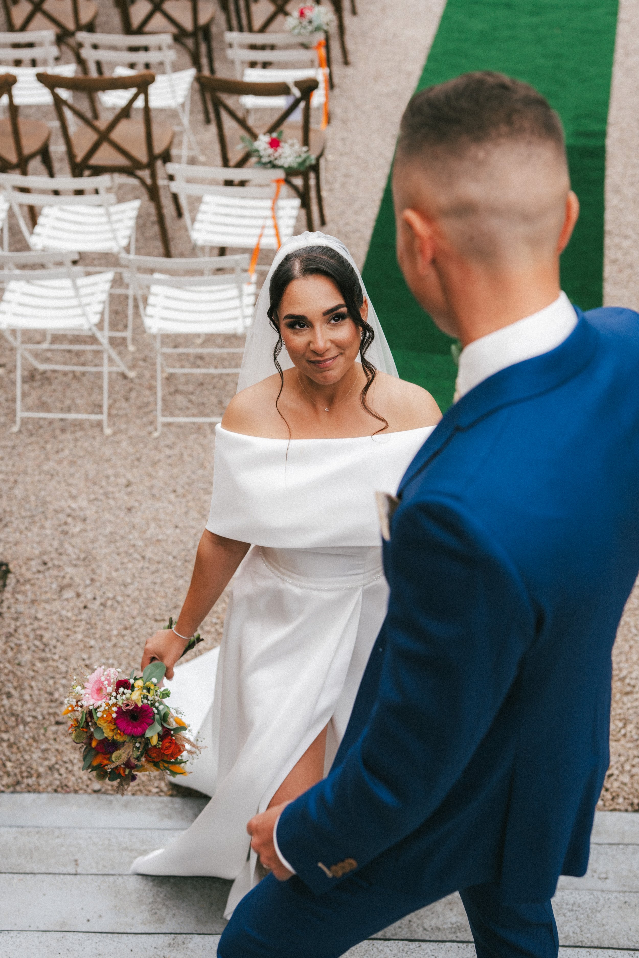Une mariée en robe de mariage blanche regarde un homme en costume bleu lors de leur mariage, avec des chaises décorées en arrière-plan.