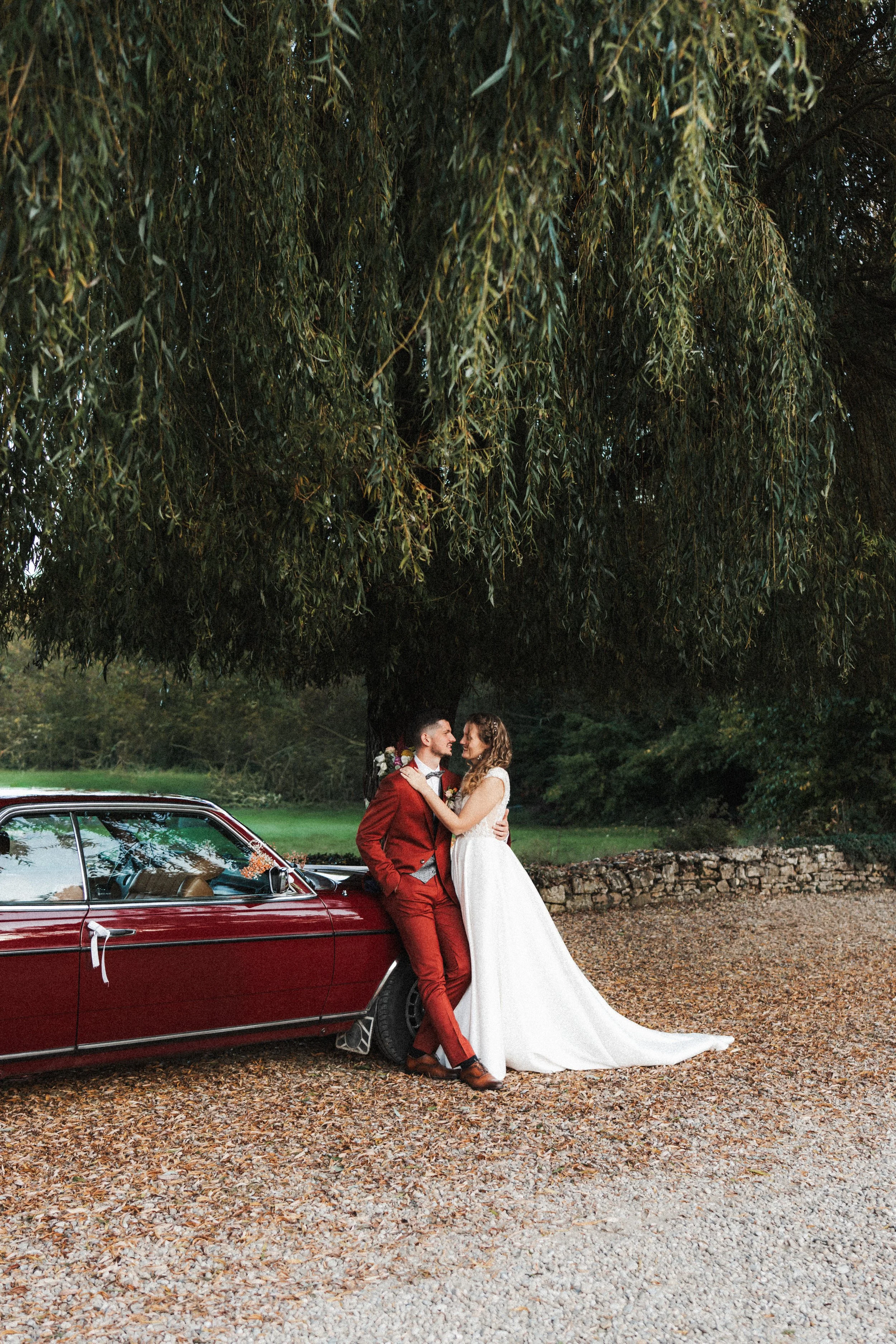 Un couple en robe de mariage posant avec une voiture ancienne sous un arbre.