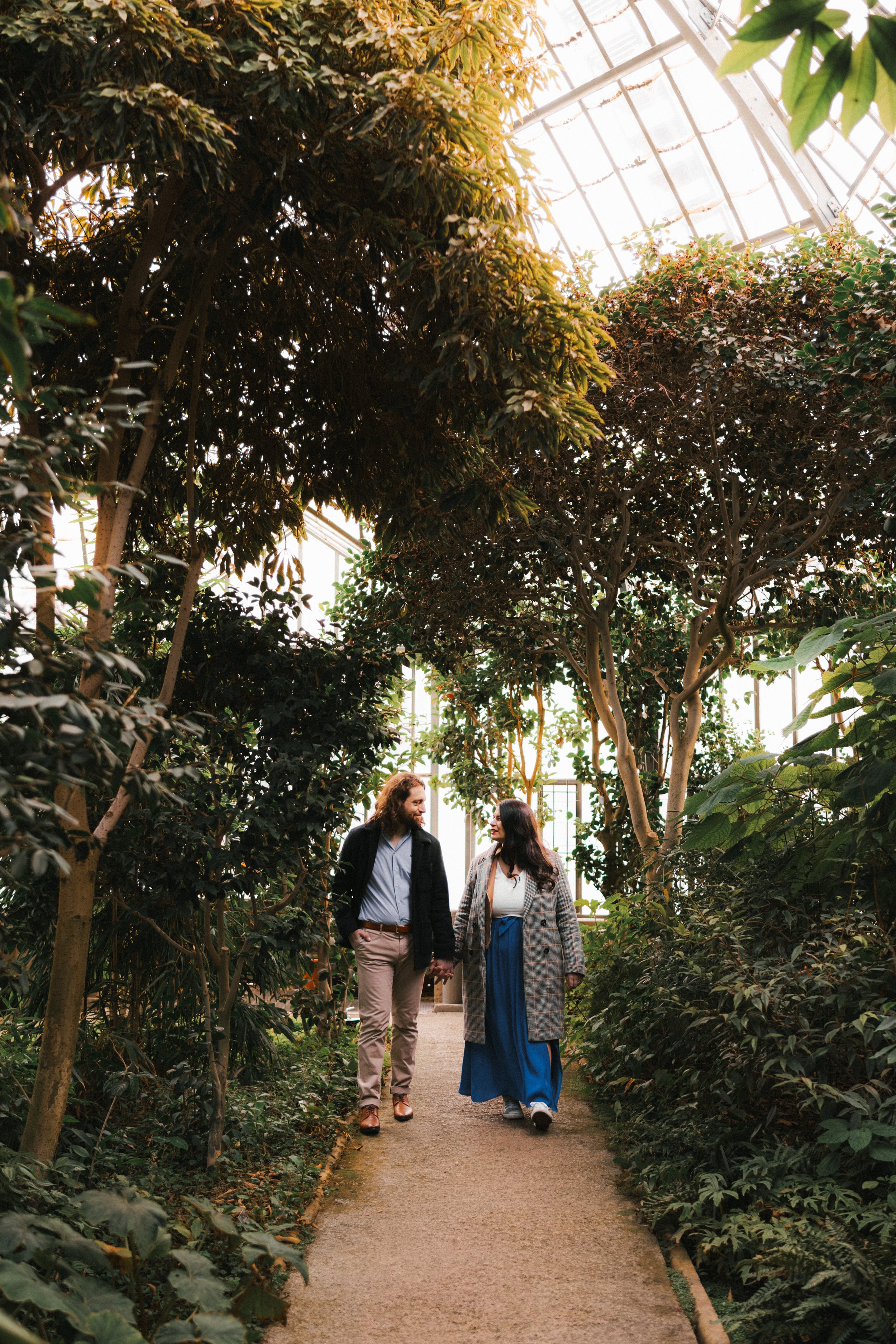 Un couple marche main dans la main à travers une serre remplie de plantes et d'arbres verts, le toit en verre laissant passer la lumière naturelle.