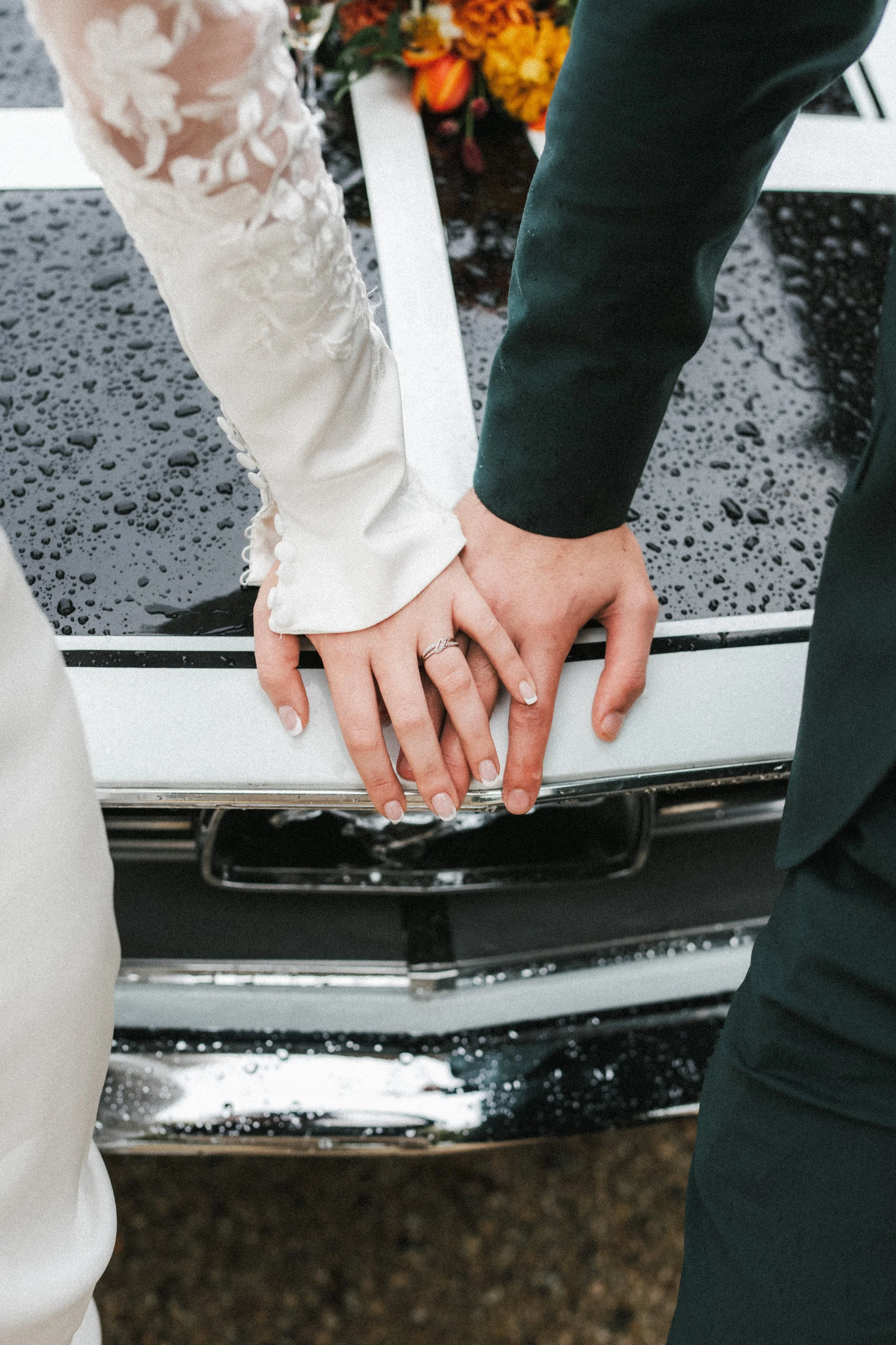 Les mains d'un couple marié, avec la mariée portant une alliance, posées sur le capot d'une voiture noire avec des gouttelettes de pluie, lors d'un mariage.