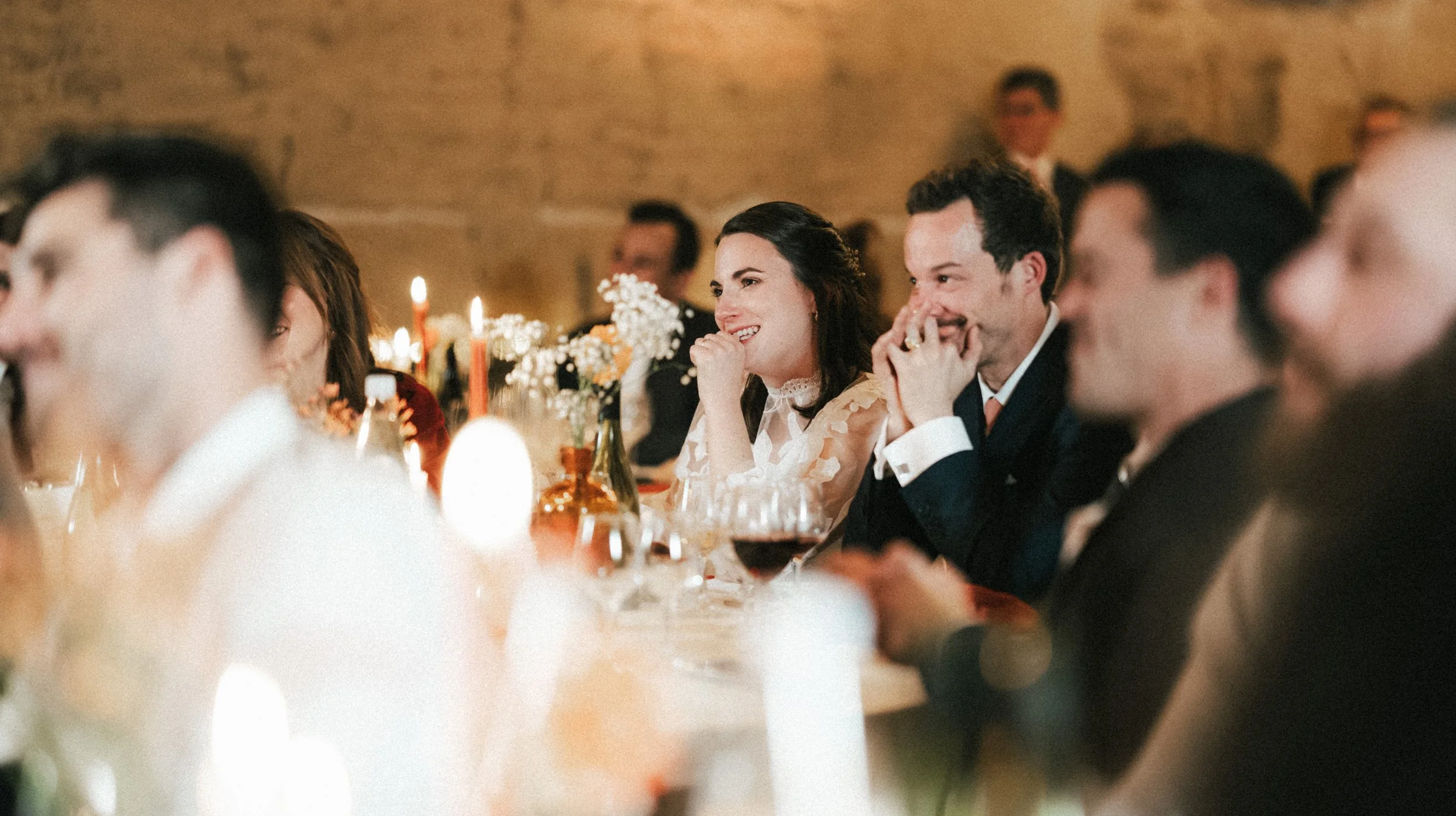 Groupe de personnes souriantes assises à une table lors d'une réception, décoration florale et bougies en fond.