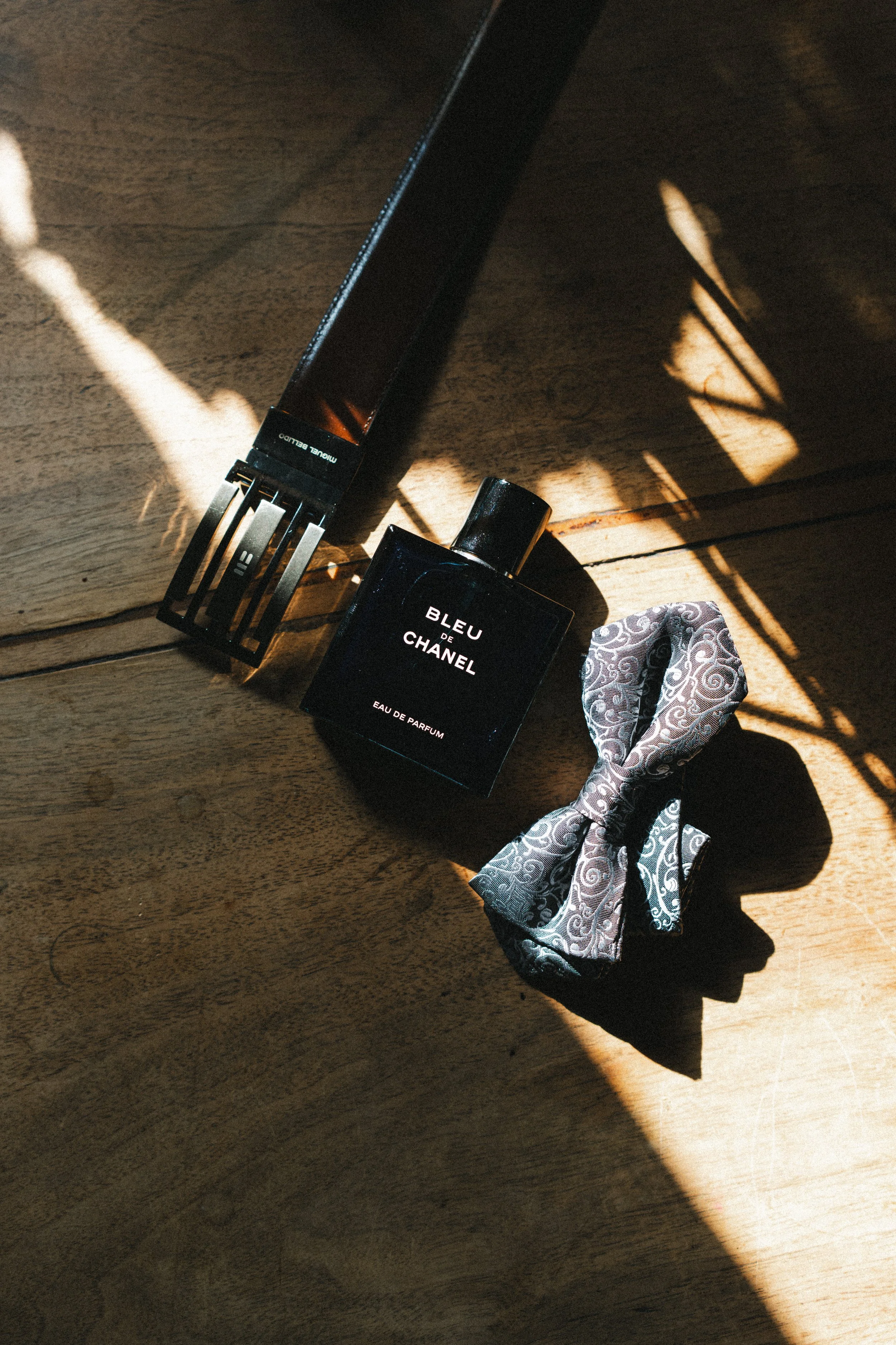 Un flacon de parfum noir 'Bleu de Chanel', une barre de cheveux en satin avec un motif de swirl, et une pince à cheveux noire sur un plan en bois avec des ombres lumineuses.