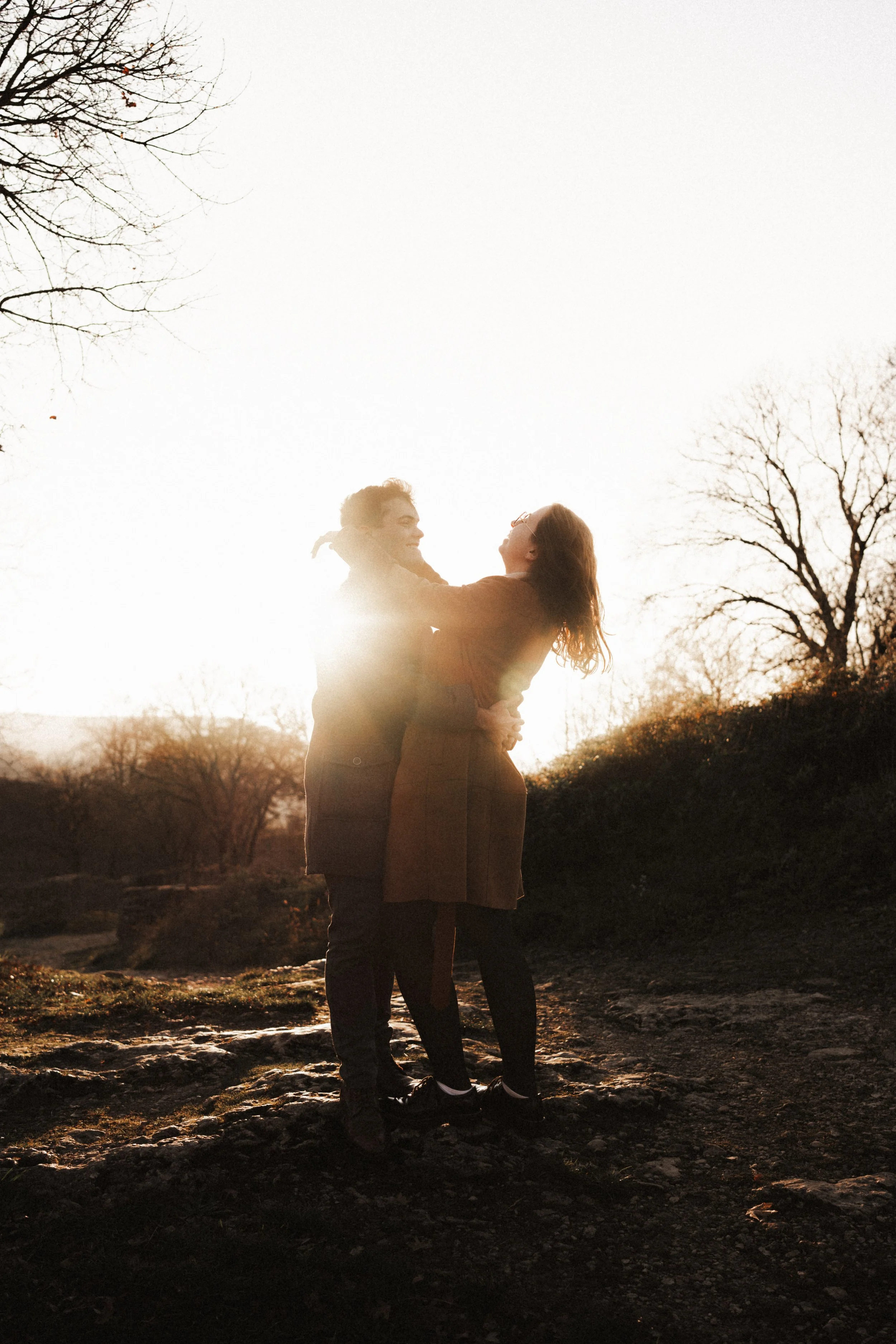Un couple souriant, enlaçant dans un paysage en plein air au coucher du soleil avec des arbres nus en arrière-plan.