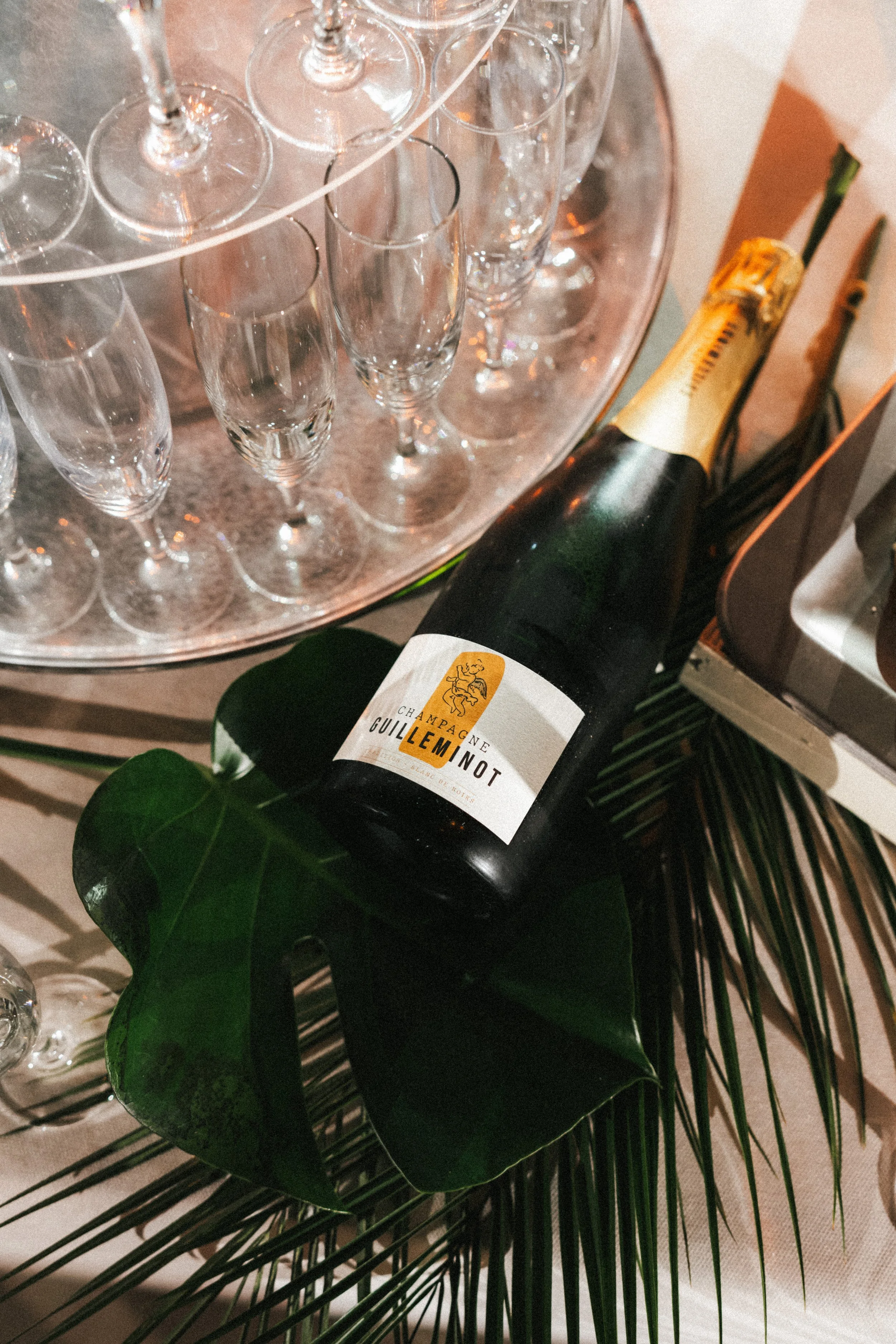 Une bouteille de champagne Gulleminot posée sur de grandes feuilles vertes, entourée de vases avec des flûtes à champagne vides sur une table décorative.