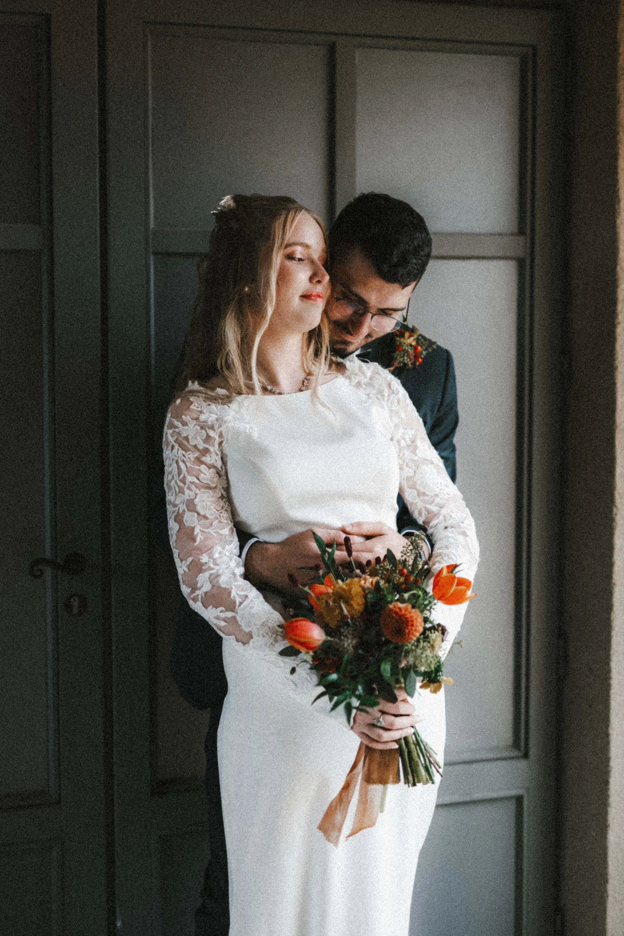 Une femme en robe de mariée tenant un bouquet de fleurs, avec un homme derrière elle, dans une ambiance intime et chaleureuse.