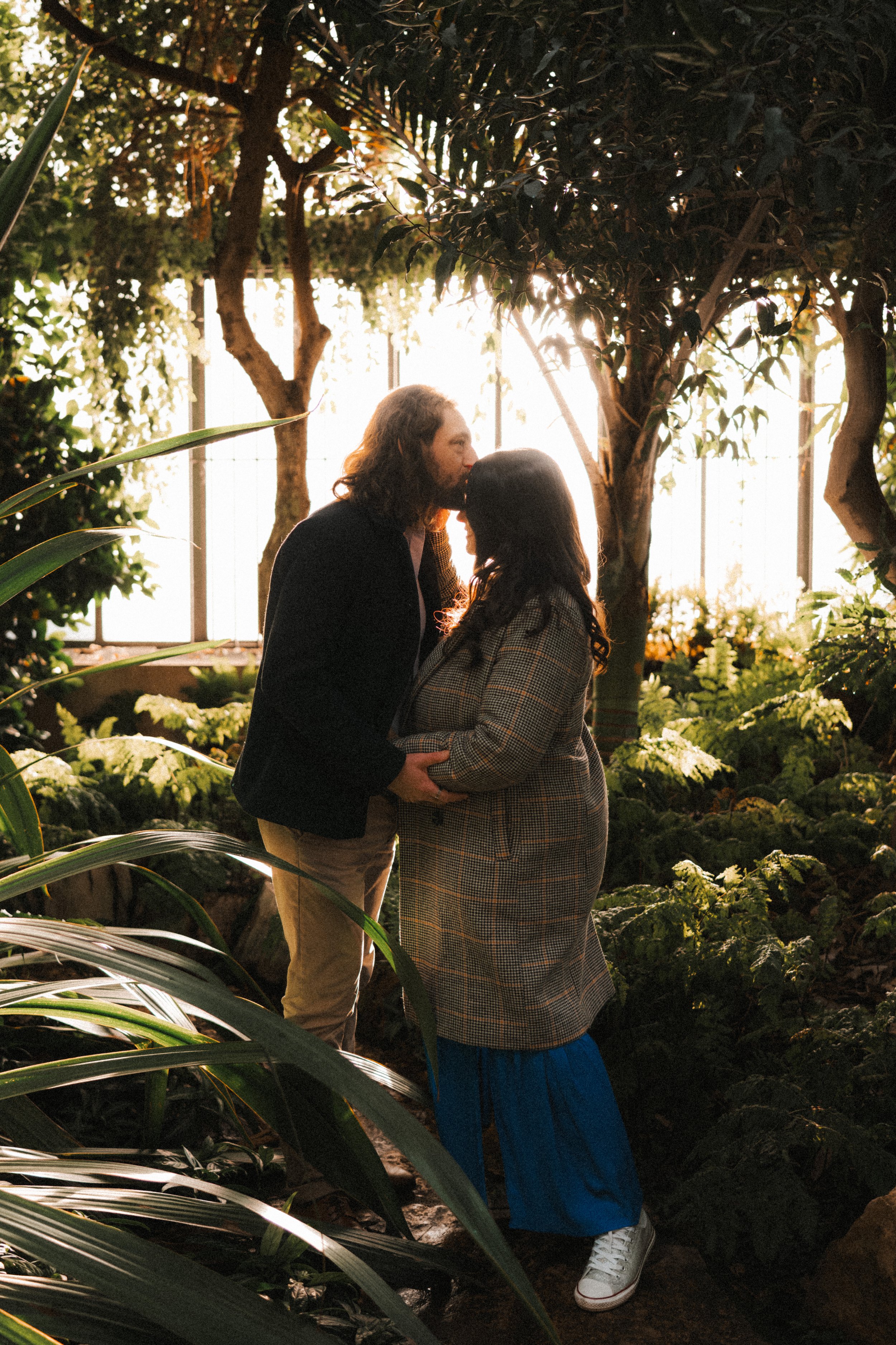 Un couple s'embrassant tendrement dans une serre ou un jardin tropical, entouré de végétation luxuriante et ensoleillée.