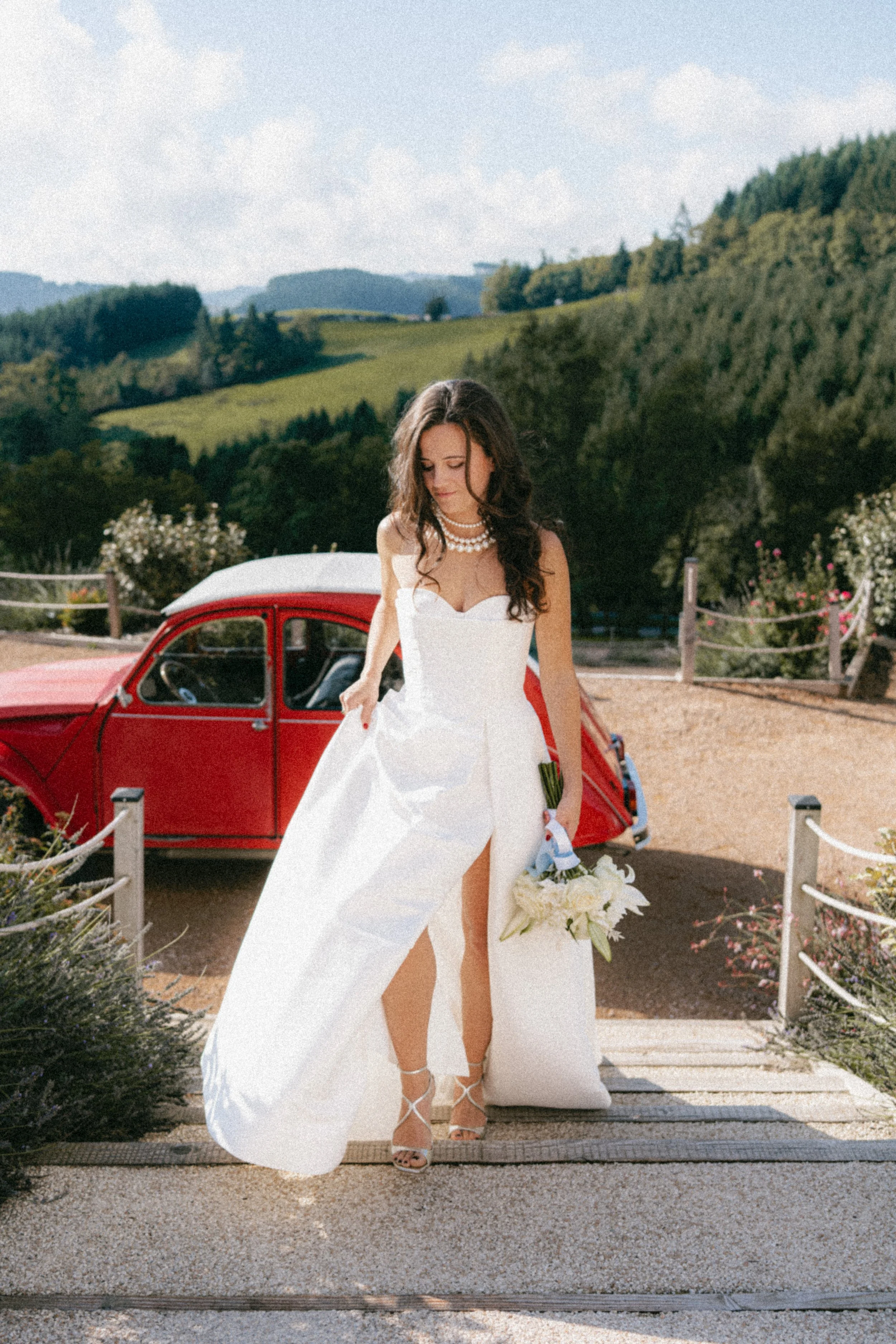 Une femme en robe de mariage blanche portant un bouquet de fleurs, devant une voiture rouge vintage, dans un paysage rural verdoyant sous un ciel bleu avec quelques nuages.
